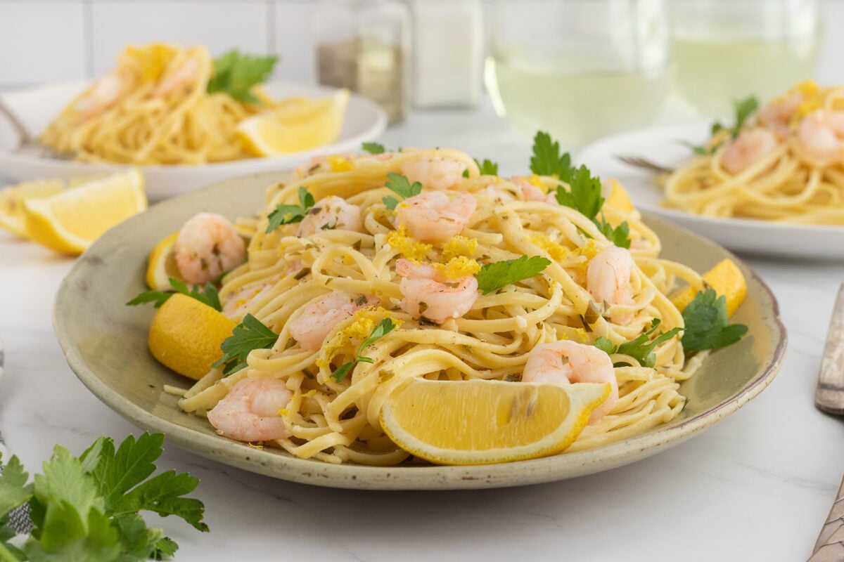 A plate of shrimp pasta garnished with fresh parsley and lemon wedges, with more pasta dishes and glasses of white wine in the background on a white table.