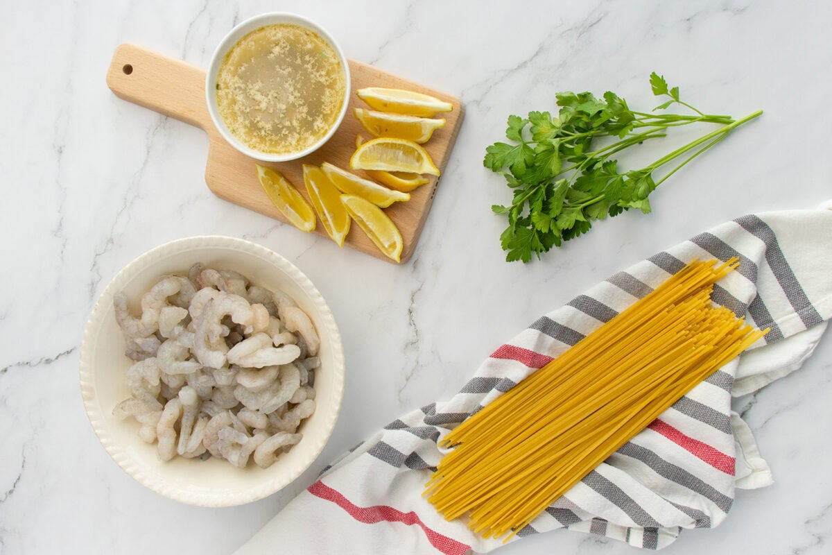 Raw shrimp in a bowl, dry spaghetti on a striped towel, lemon wedges and a bowl of broth on a wooden board, and fresh parsley on a marble surface.