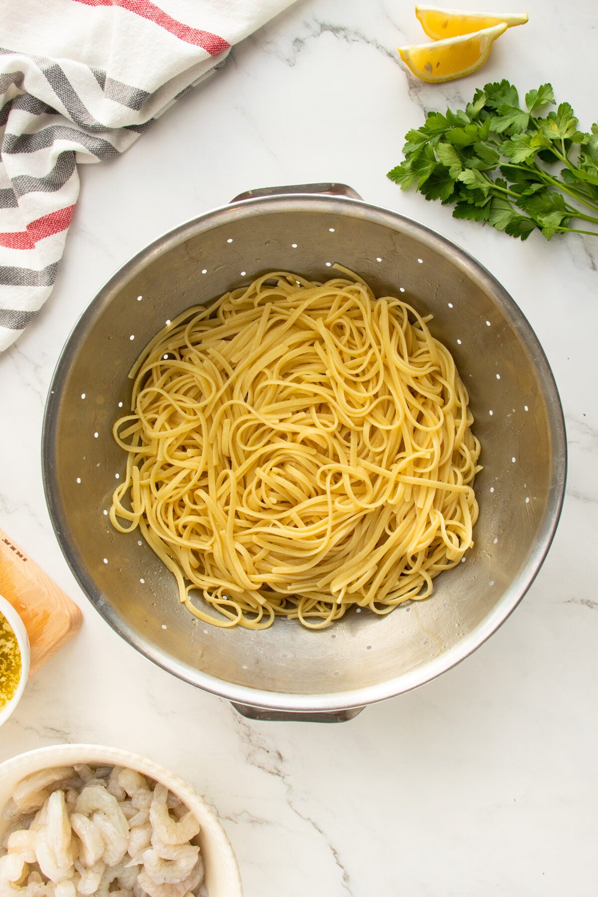 Cooked spaghetti noodles are draining in a metal colander on a marble countertop. Surrounding the colander are fresh parsley, lemon wedges, a striped kitchen towel, a bowl of shrimp, and a container of grated cheese.