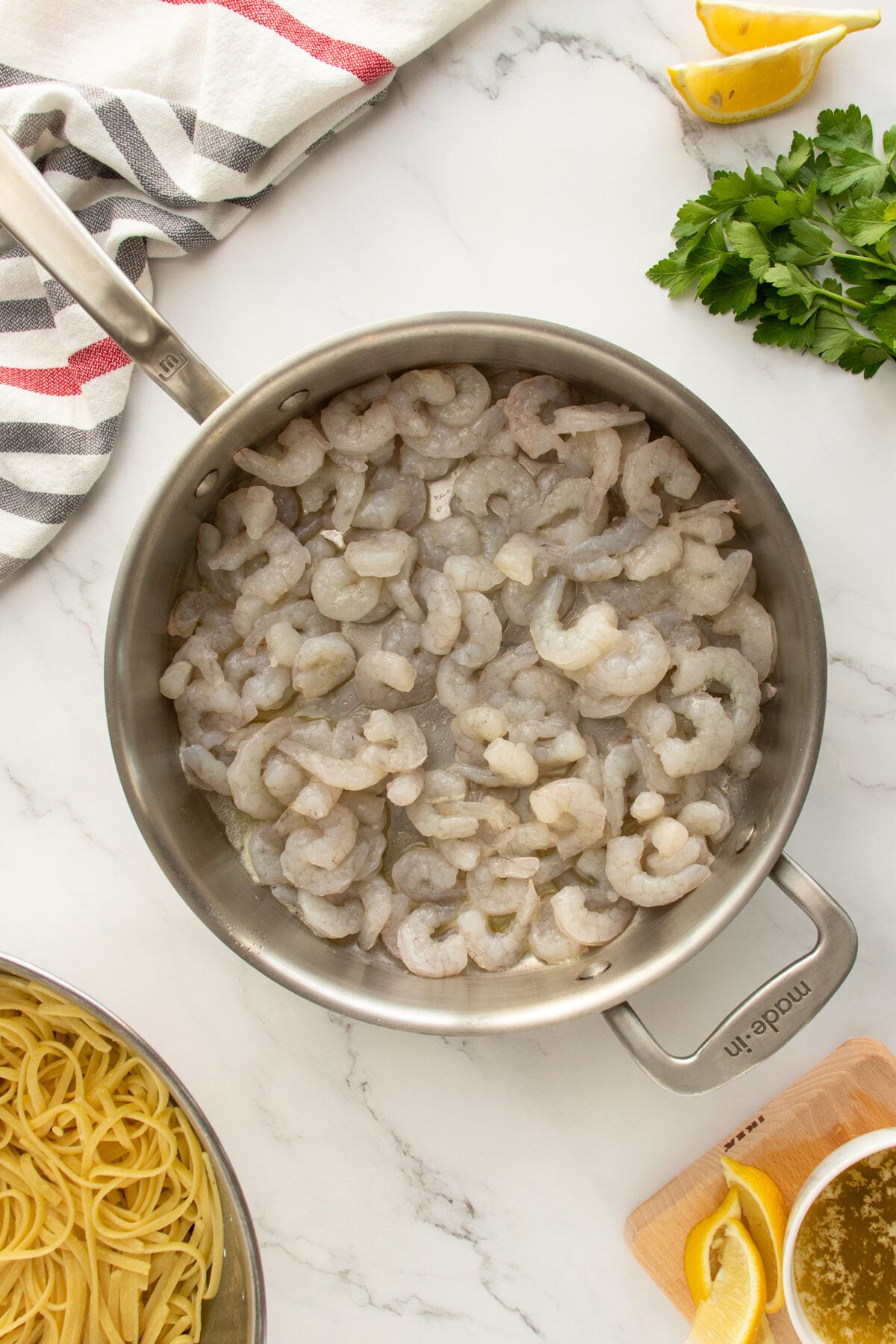 A stainless steel pan filled with raw shrimp sits on a marble countertop. Nearby are uncooked pasta, lemon wedges, fresh parsley, a small bowl of sauce, and a striped kitchen towel.