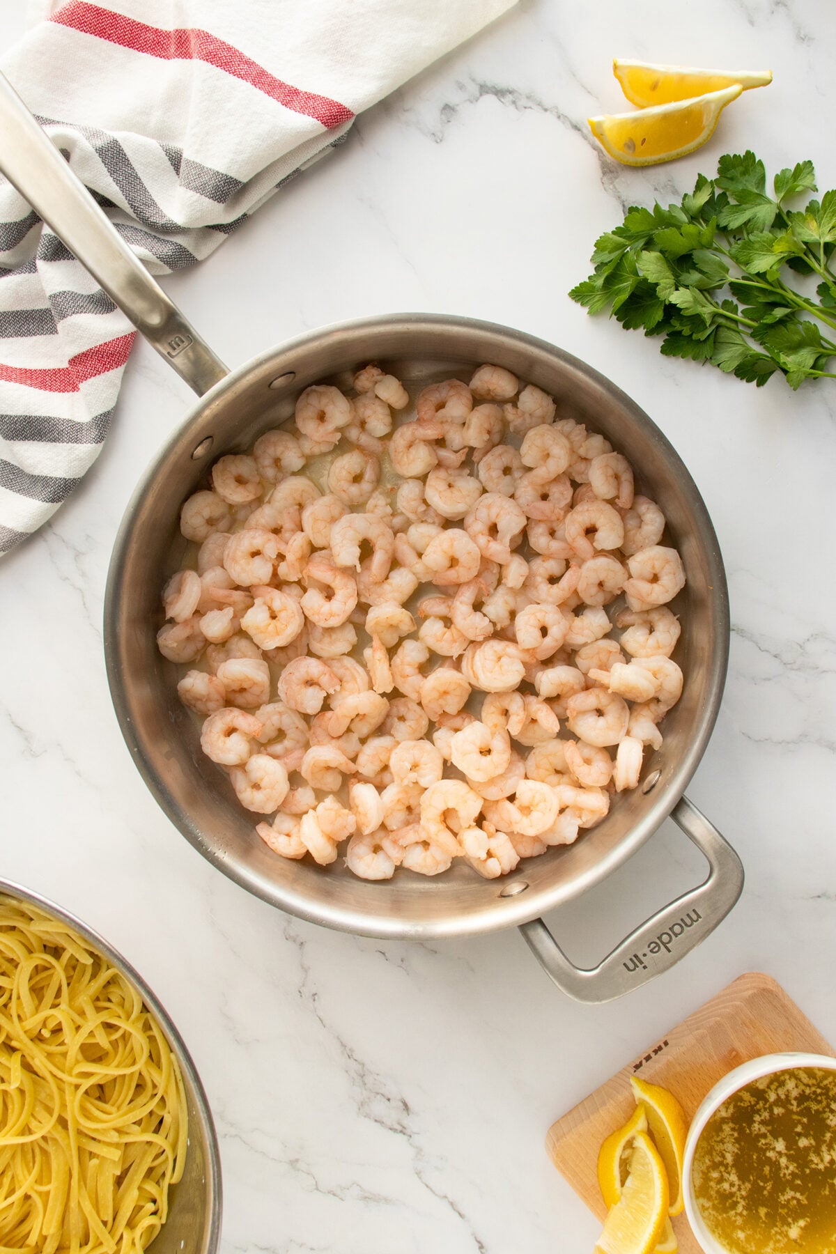 A stainless steel pan filled with cooked shrimp sits on a white marble countertop. Nearby are a bowl of pasta, lemon wedges, fresh parsley, a striped towel, and a small bowl of sauce.