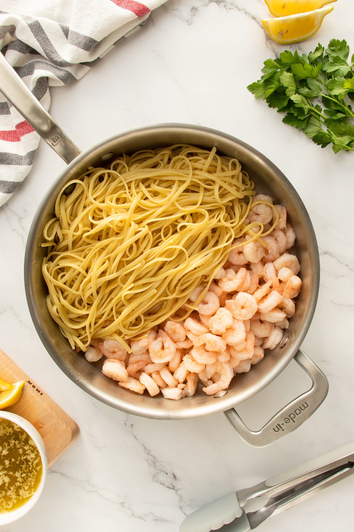 A stainless steel pan containing cooked linguine pasta and shrimp sits on a marble countertop, surrounded by lemon wedges, fresh parsley, a striped towel, tongs, and a bowl of sauce.