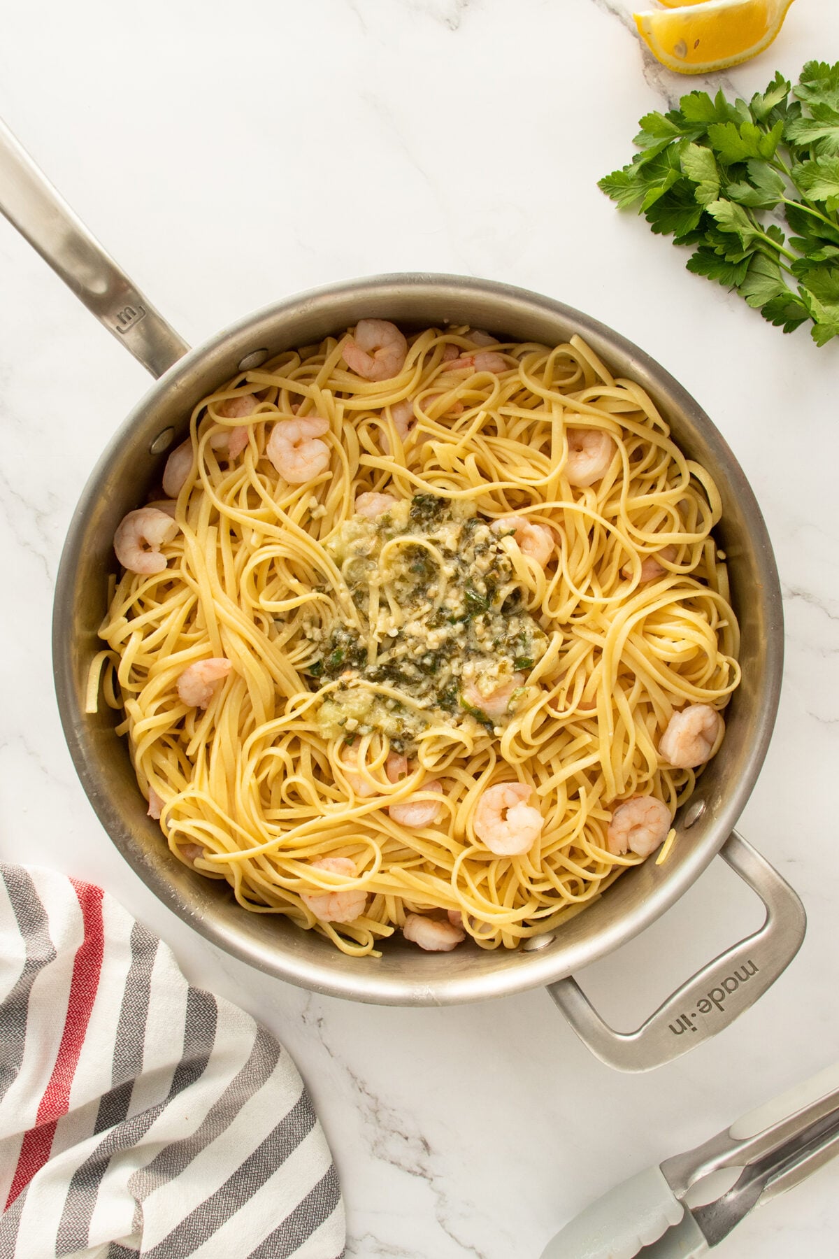 A skillet filled with cooked linguine pasta, shrimp, and a creamy sauce in the center. Fresh parsley, a lemon wedge, and a striped kitchen towel are nearby on a marble countertop.