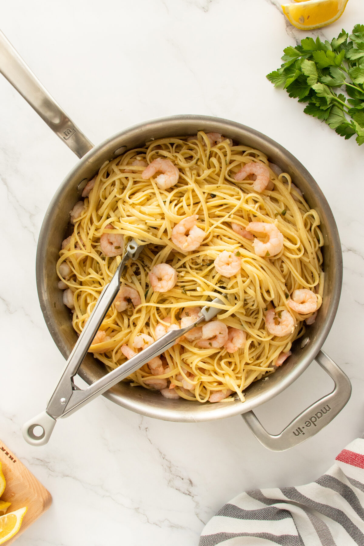 A stainless steel pan filled with cooked spaghetti and shrimp sits on a marble counter, with metal tongs resting inside. Fresh parsley, lemon wedges, and a striped kitchen towel are nearby.