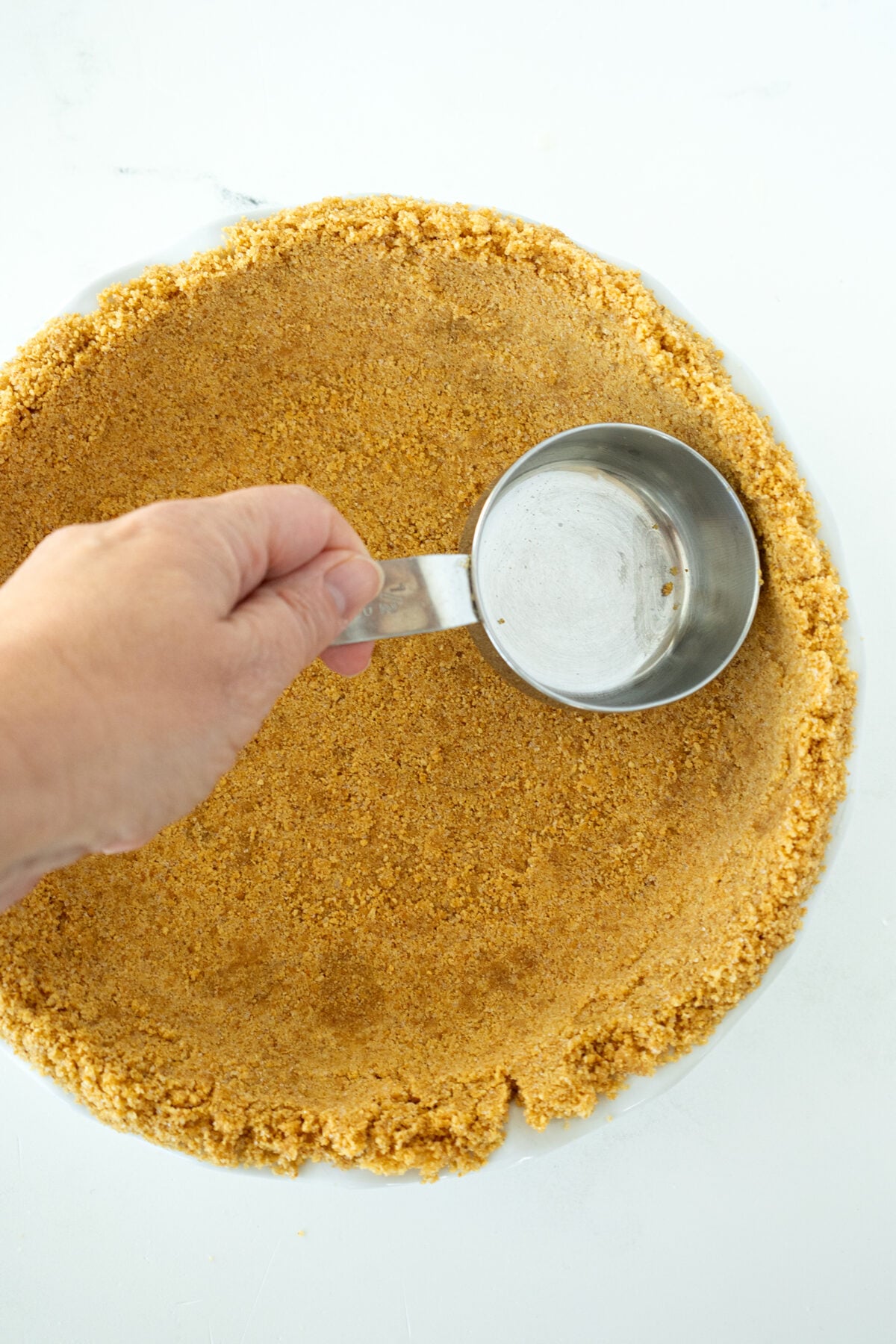 A hand presses a metal measuring cup into a pie dish filled with a crumbly graham cracker crust, shaping the crust evenly along the bottom and edges.
