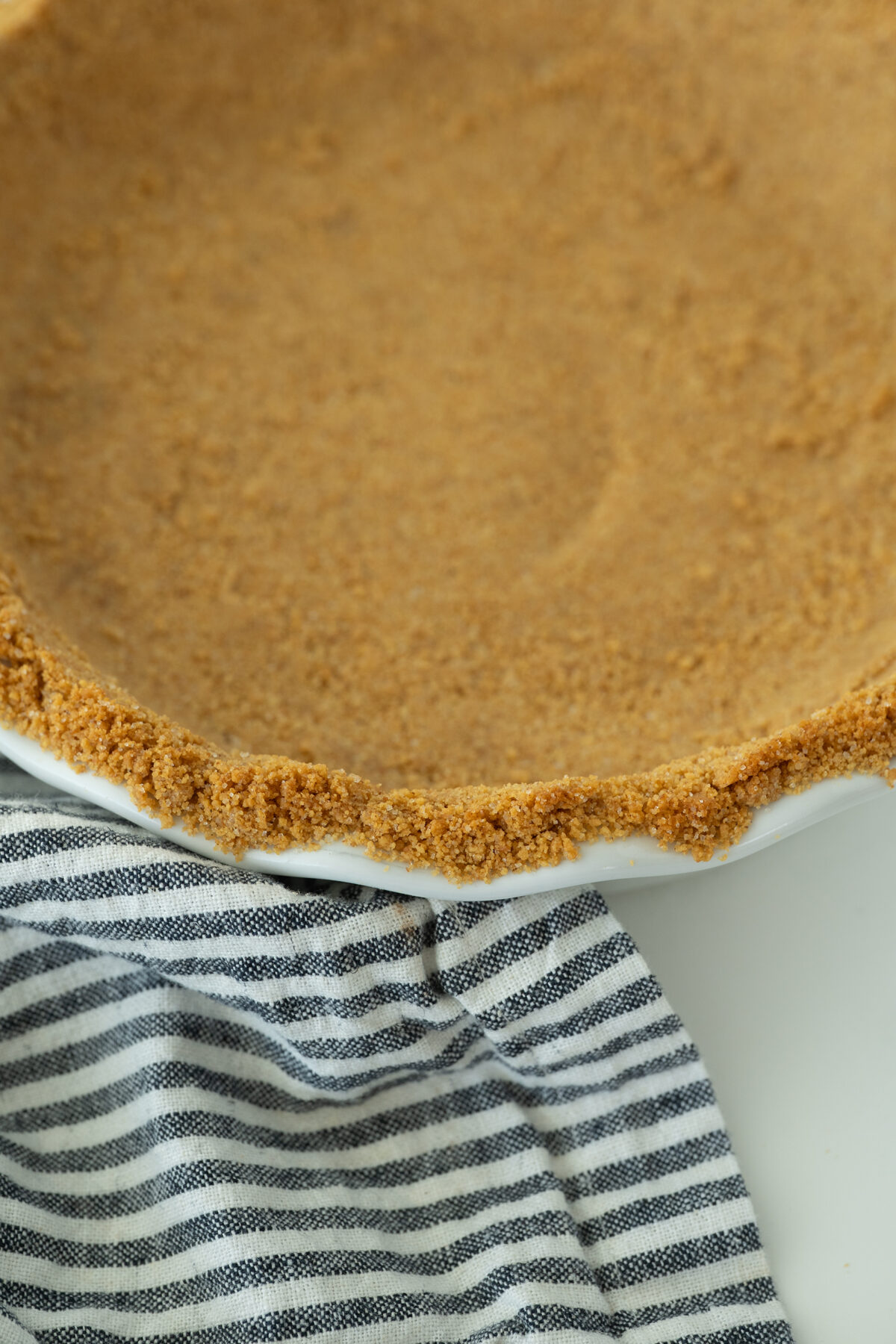 A close-up of a pie dish with a pressed graham cracker crust, placed next to a black and white striped kitchen towel.