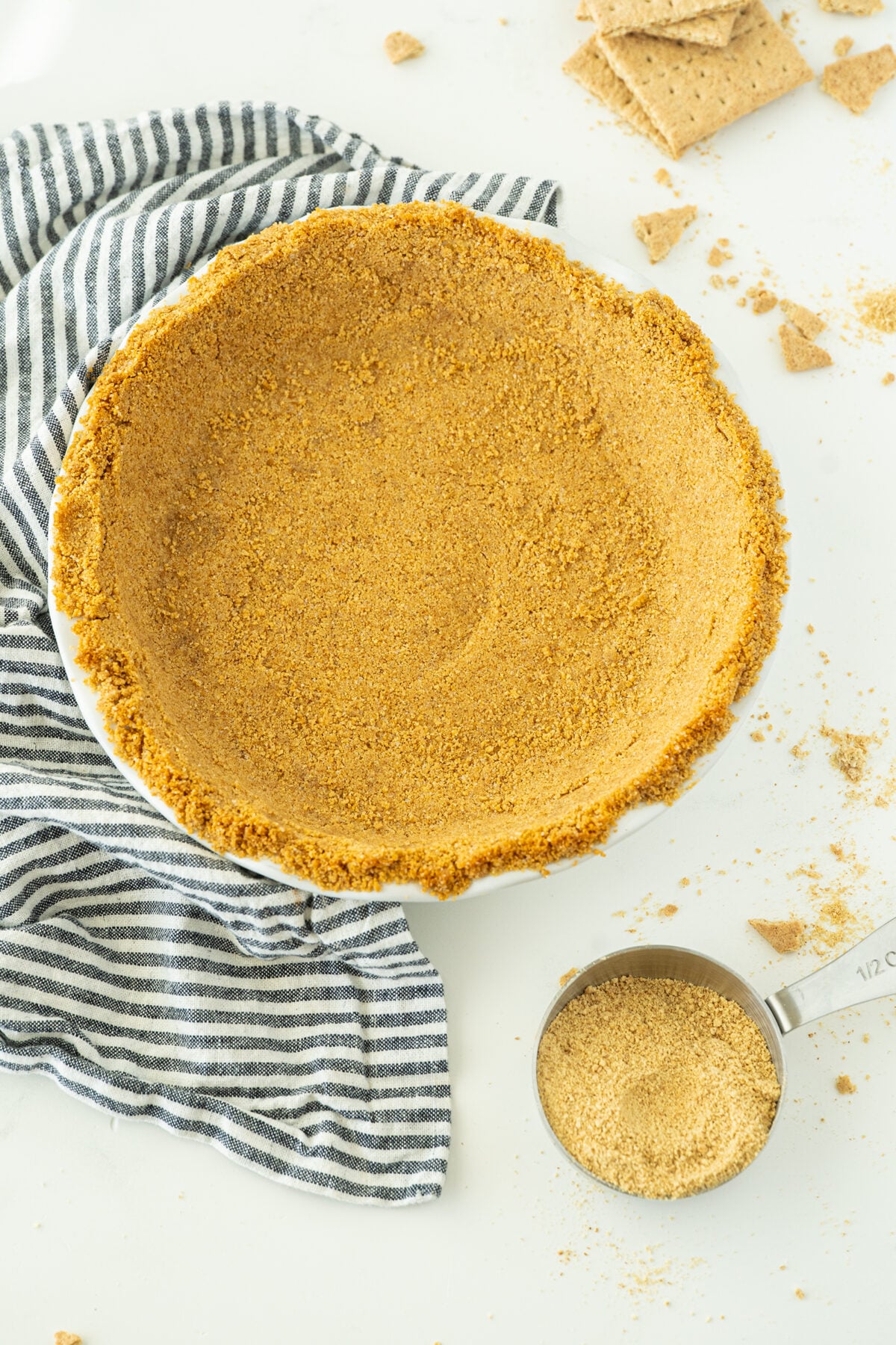 A pie dish filled with a graham cracker crust sits on a white surface next to a striped cloth, crushed graham crackers, and a measuring cup containing more graham cracker crumbs.