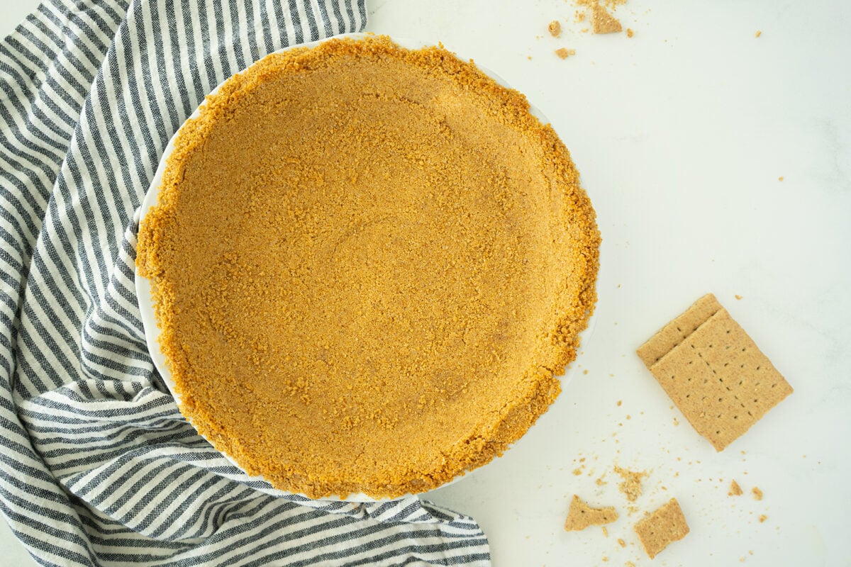 A pie dish filled with a graham cracker crust sits on a white surface beside a striped cloth, with graham cracker pieces and crumbs scattered nearby.
