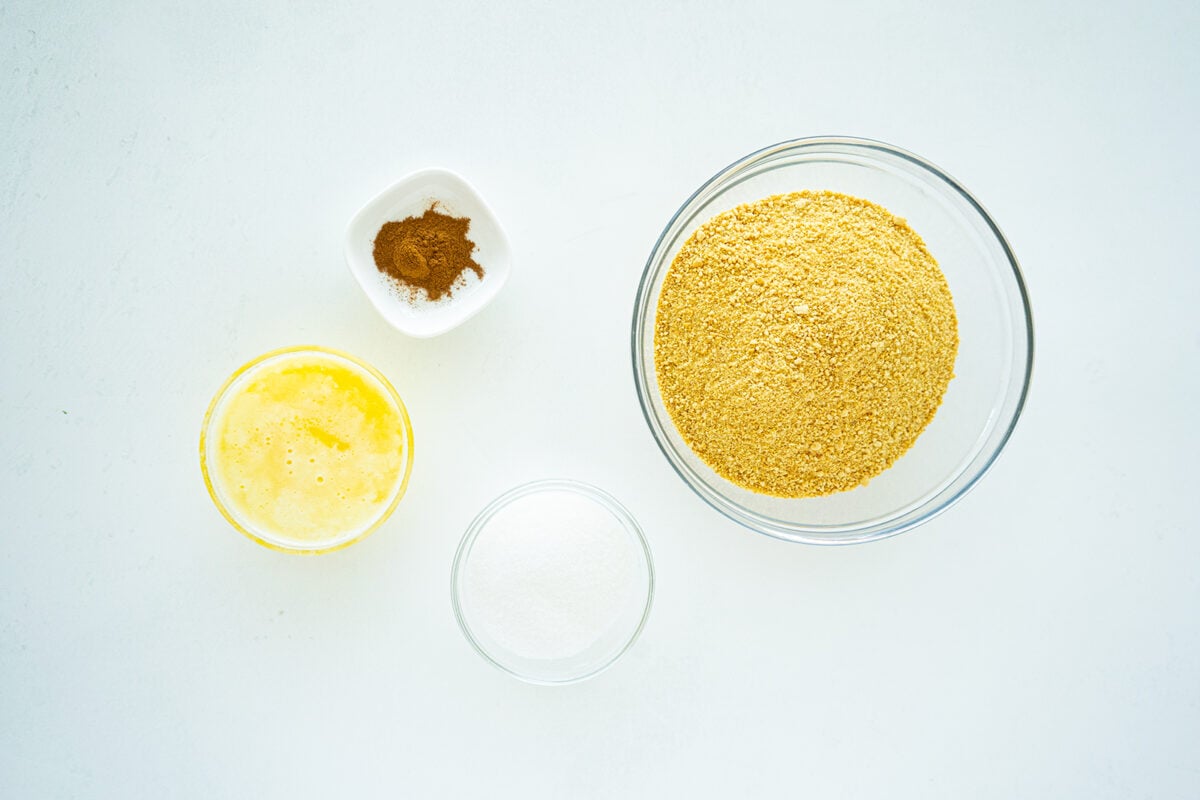 Four ingredients arranged on a white surface: a bowl of graham cracker crumbs, a bowl of sugar, a bowl of melted butter, and a small dish with a mixture of cinnamon and salt.