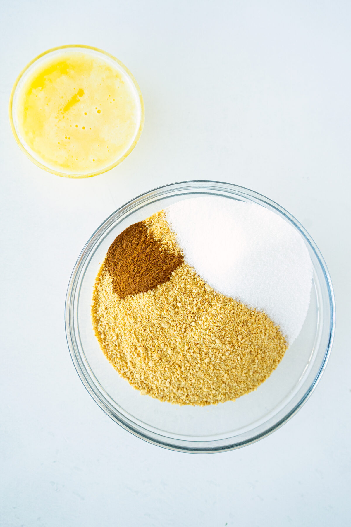 A glass bowl containing granulated sugar, ground cinnamon, and graham cracker crumbs sits next to a smaller bowl of melted butter on a white surface.