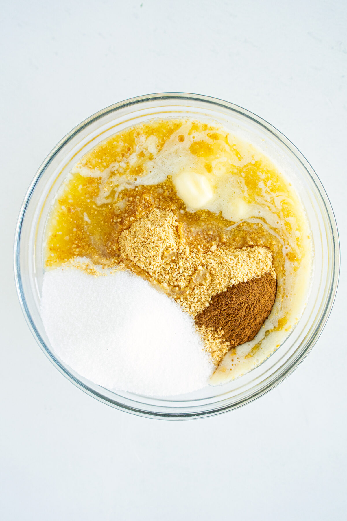 A glass bowl containing melted butter, white sugar, brown sugar, cinnamon, and other spices on a white surface, seen from above.