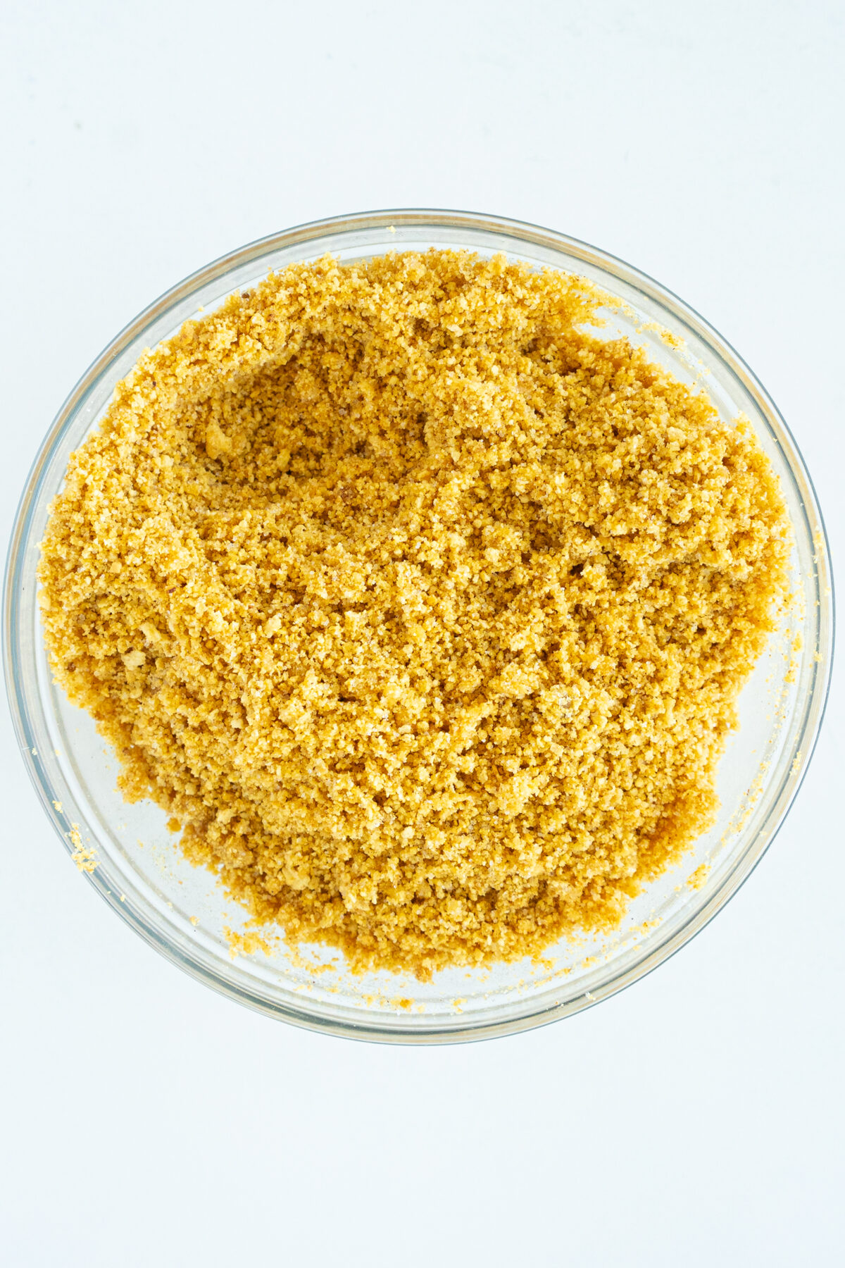 A clear glass bowl filled with golden-yellow, finely ground breadcrumb mixture, viewed from above on a white background.