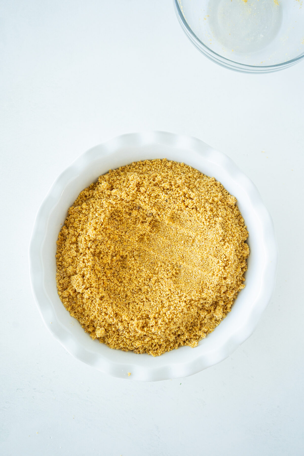 A white pie dish filled with finely ground yellow graham cracker crumbs sits on a white surface, with part of a clear glass bowl visible at the top right corner.