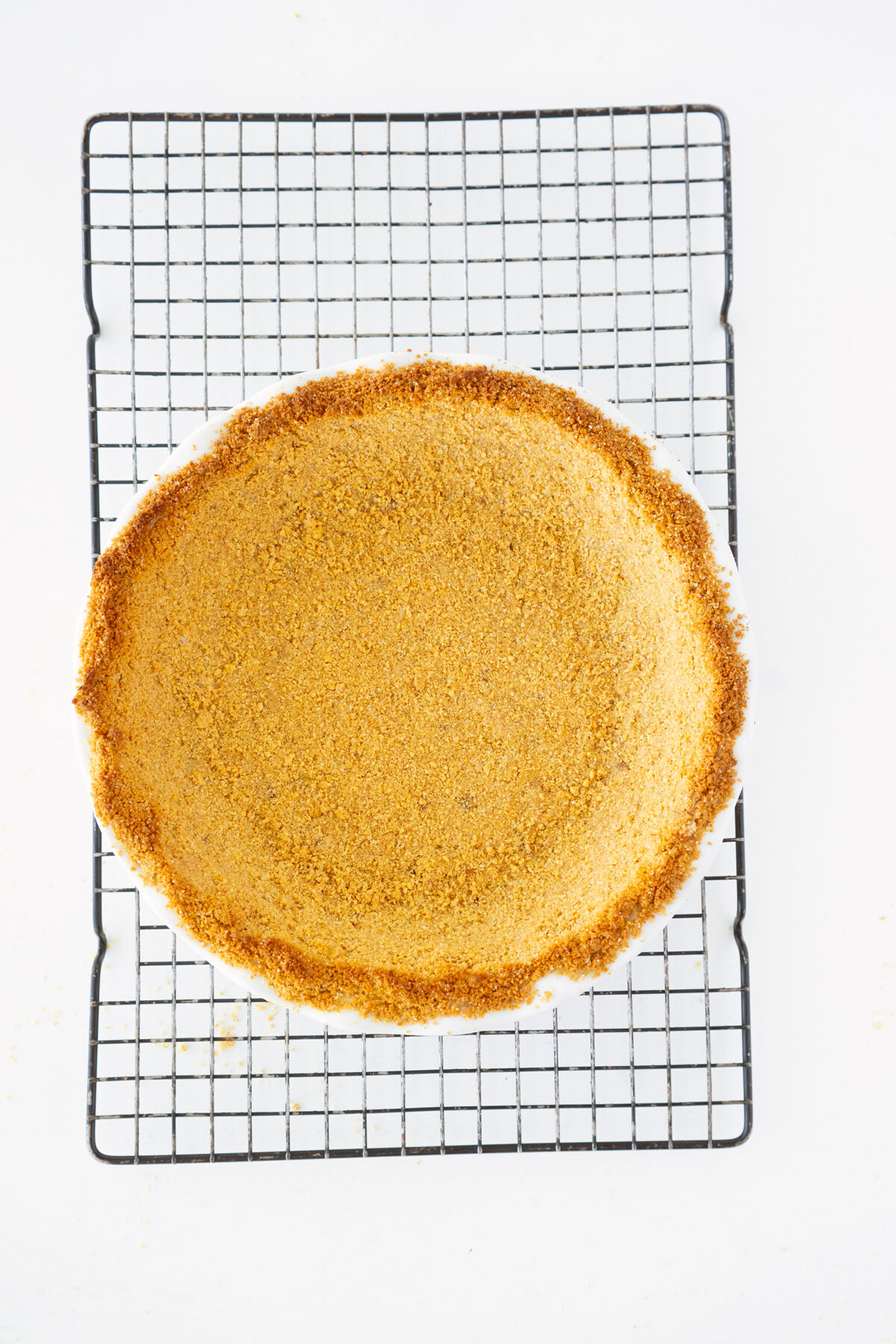 A baked graham cracker pie crust sits on a black cooling rack against a white background.