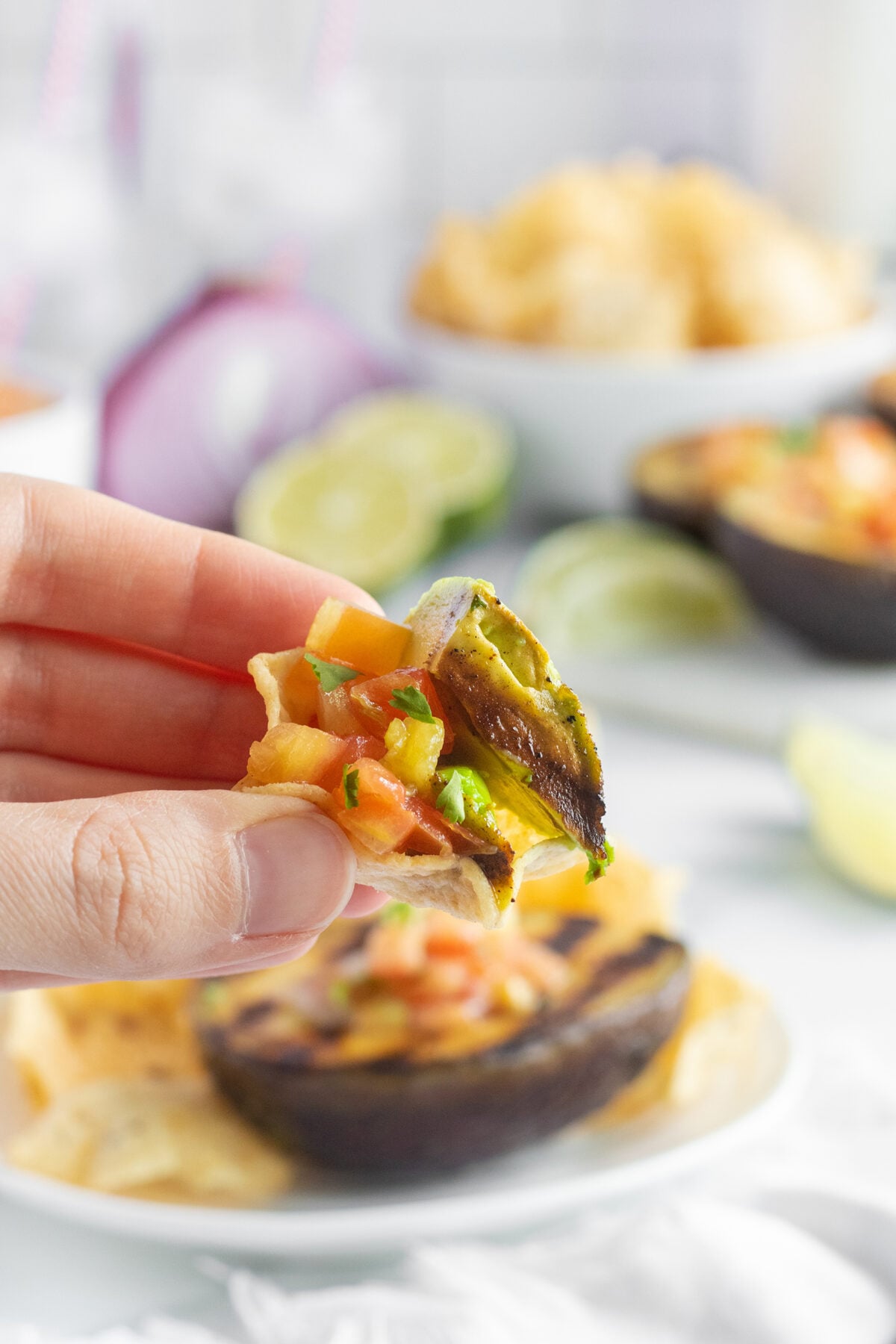 A hand holds a tortilla chip topped with sliced grilled avocado, diced tomatoes, onions, and jalapeños. In the background, there are lime slices, chips, and a halved avocado on a plate.