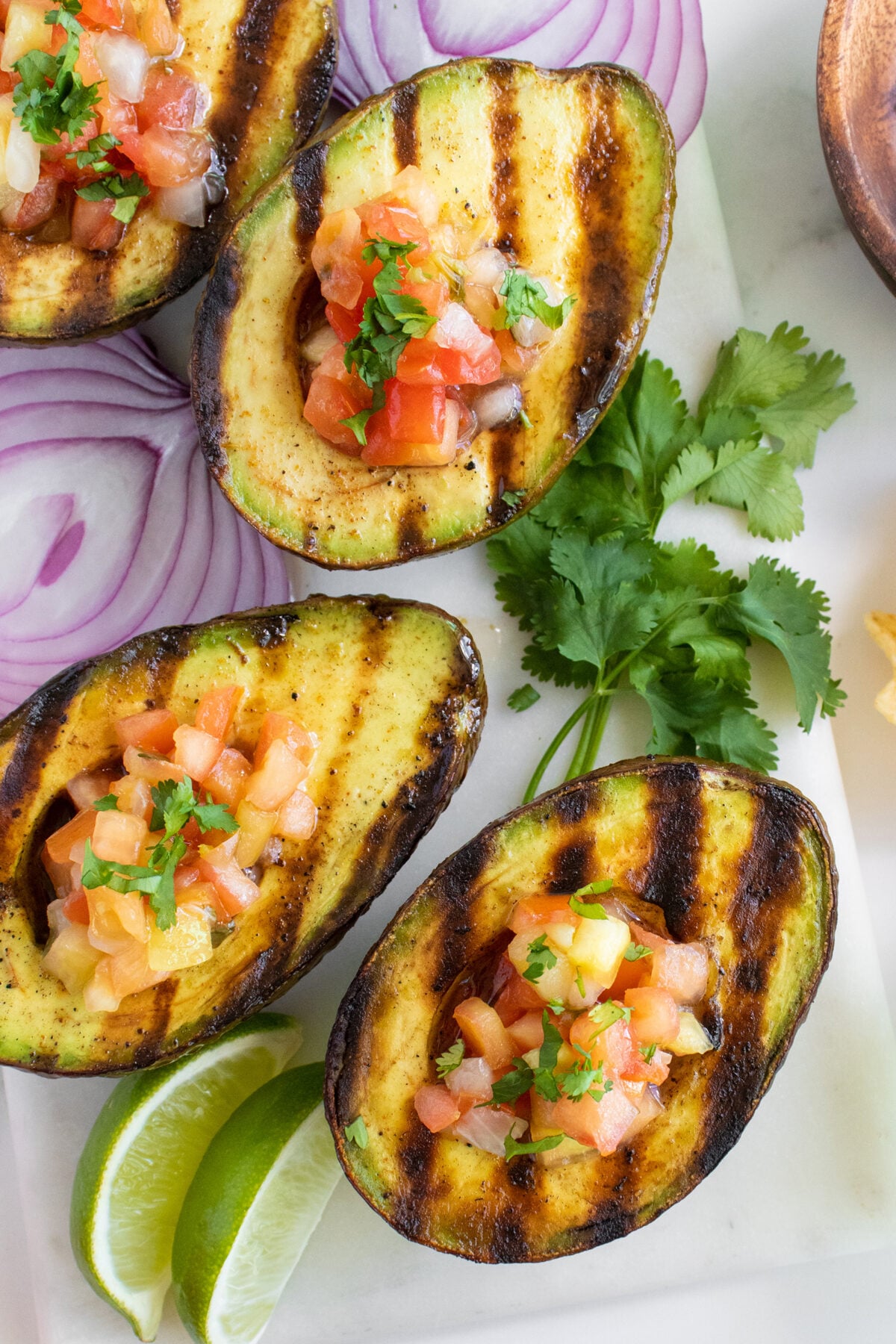 Grilled avocado halves filled with fresh salsa, garnished with chopped cilantro, are arranged on a white surface with red onion slices, cilantro sprigs, and lime wedges nearby.