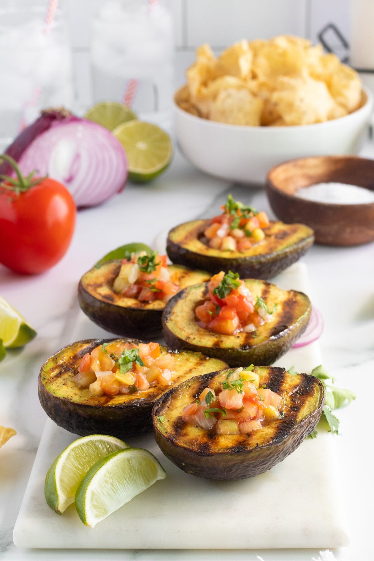 Five grilled avocado halves filled with salsa are arranged on a marble board with lime wedges. In the background are tortilla chips, a red onion, a tomato, and a cut lime.