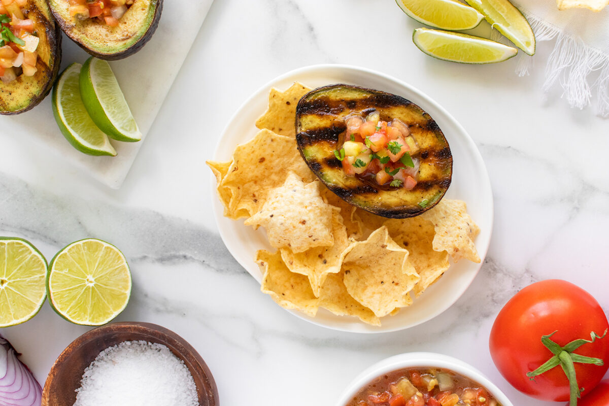 A plate of tortilla chips with a grilled avocado half filled with salsa, surrounded by lime wedges, sliced onions, a tomato, and a bowl of salsa on a marble surface.