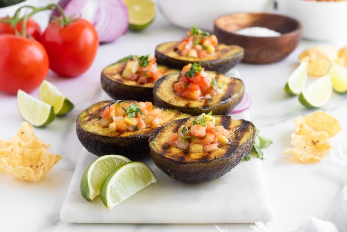 Grilled avocado halves filled with fresh salsa, garnished with cilantro, served with lime wedges on a marble board. Tomatoes, onions, tortilla chips, and a bowl of salt are in the background.