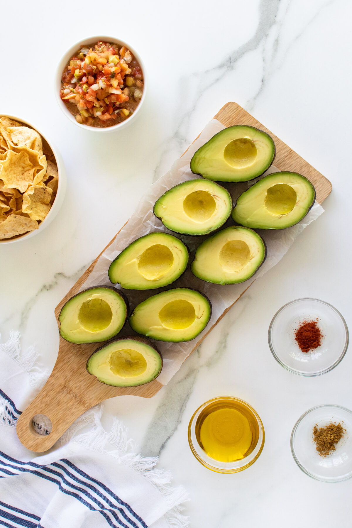 A wooden board with eight halved avocados, bowls of salsa, tortilla chips, olive oil, spices, and a striped cloth napkin on a white marble surface.