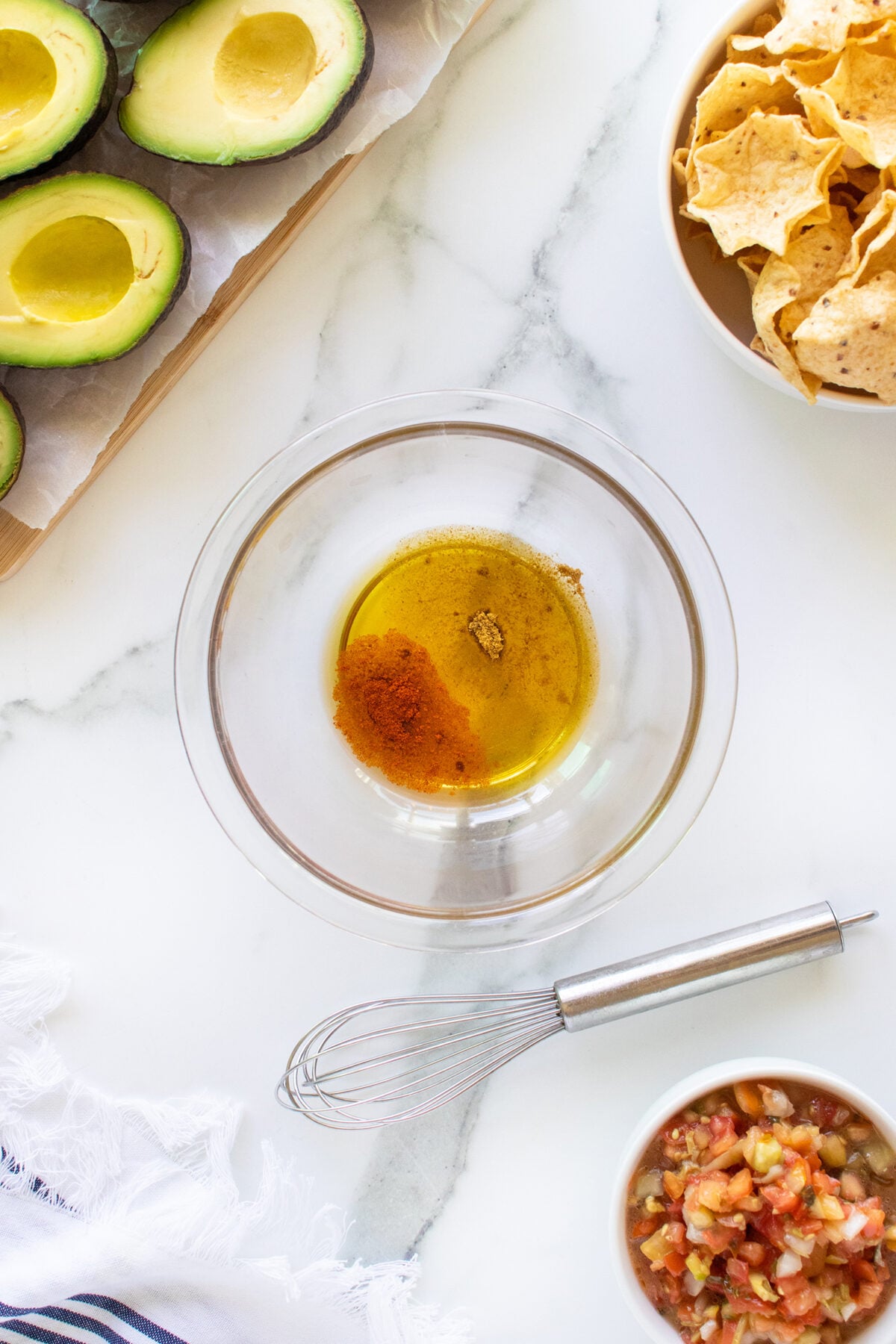 Top-down view of a glass bowl with spices and oil, surrounded by avocado halves, tortilla chips, salsa, and a whisk on a white marble surface.