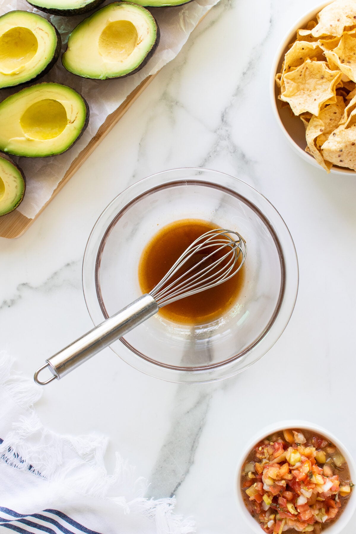 Overhead view of a glass bowl with whisk and brown liquid, surrounded by halved avocados, tortilla chips, and a bowl of salsa on a white marble surface.