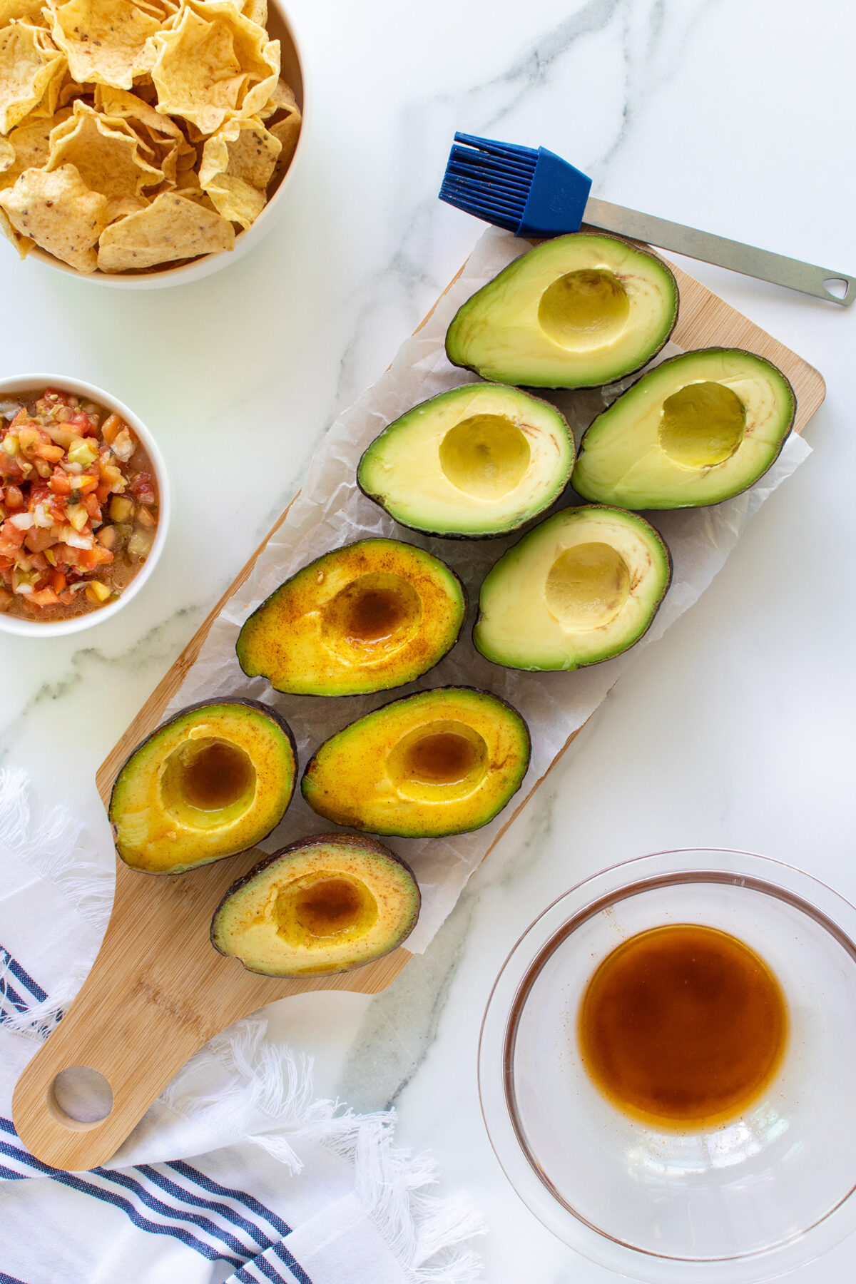 Halved avocados on a wooden board, some brushed with marinade. Nearby are a bowl of tortilla chips, a bowl of salsa, a basting brush, and a glass bowl with marinade on a white marble surface.