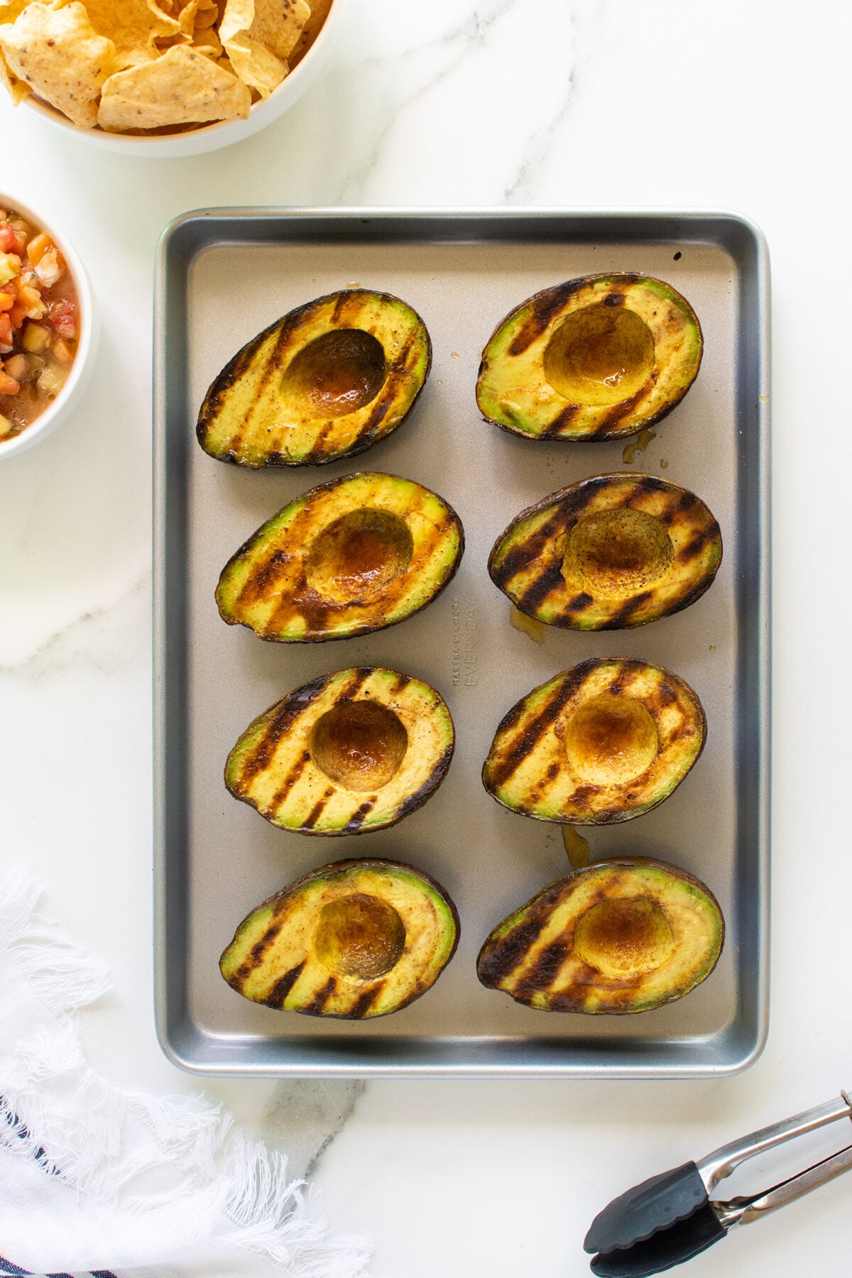 A baking pan with eight avocado halves displaying grill marks sits on a white surface. Nearby are a bowl of tortilla chips, a bowl of salsa, a white napkin, and a pair of tongs.