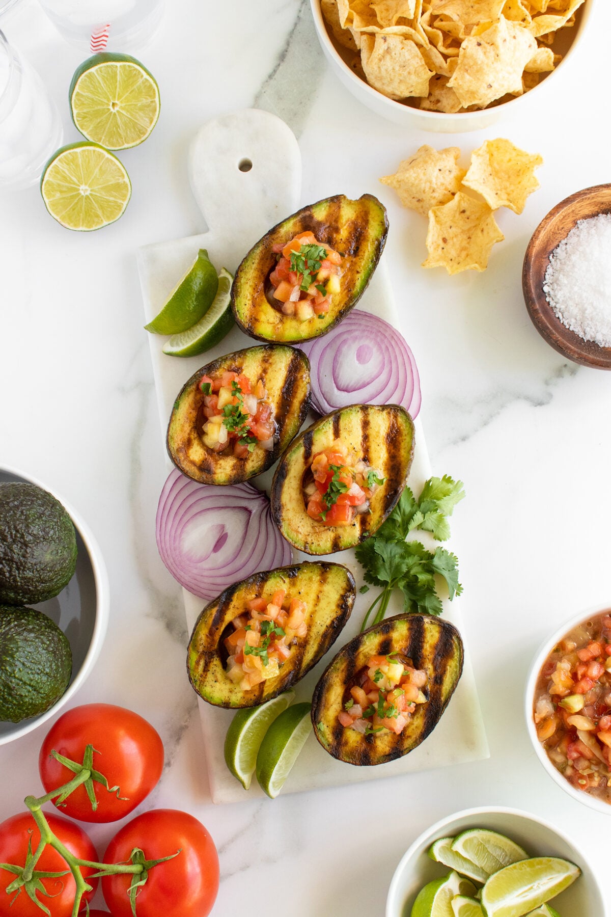Grilled avocado halves filled with salsa are arranged on a white marble board with lime wedges, red onion slices, and cilantro. Surrounding the board are bowls of chips, salt, salsa, avocados, tomatoes, and lime halves.