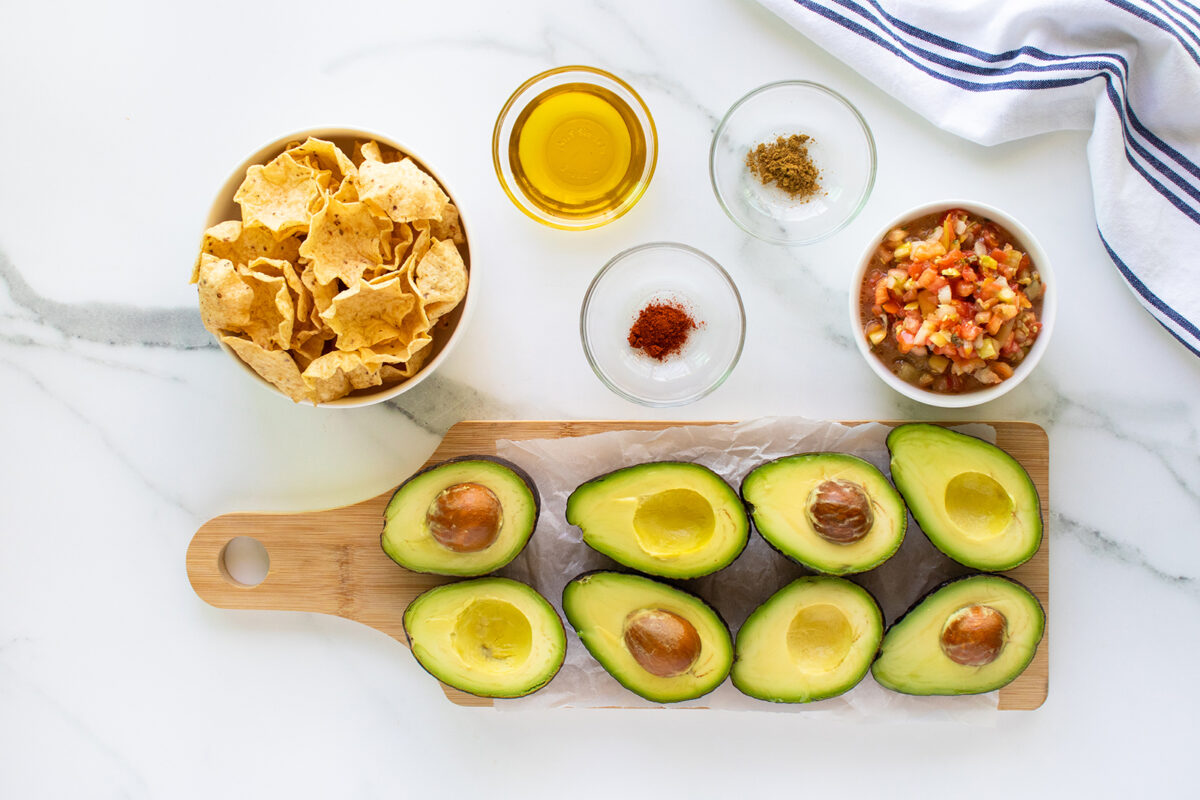 A top view of halved avocados on a wooden board, surrounded by bowls of tortilla chips, oil, spices, salsa, and a blue-striped towel on a white marble surface.