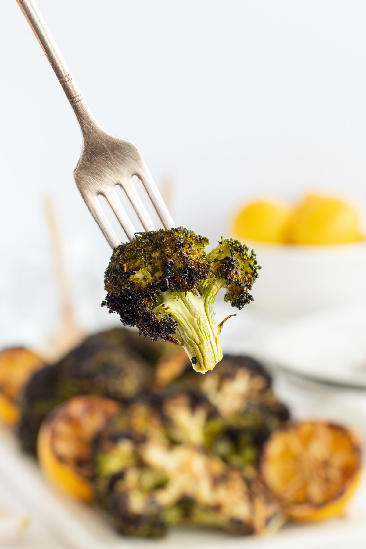 A close-up of a fork holding a piece of roasted broccoli, with charred edges. In the blurred background, more roasted broccoli and sliced lemons are on a plate.