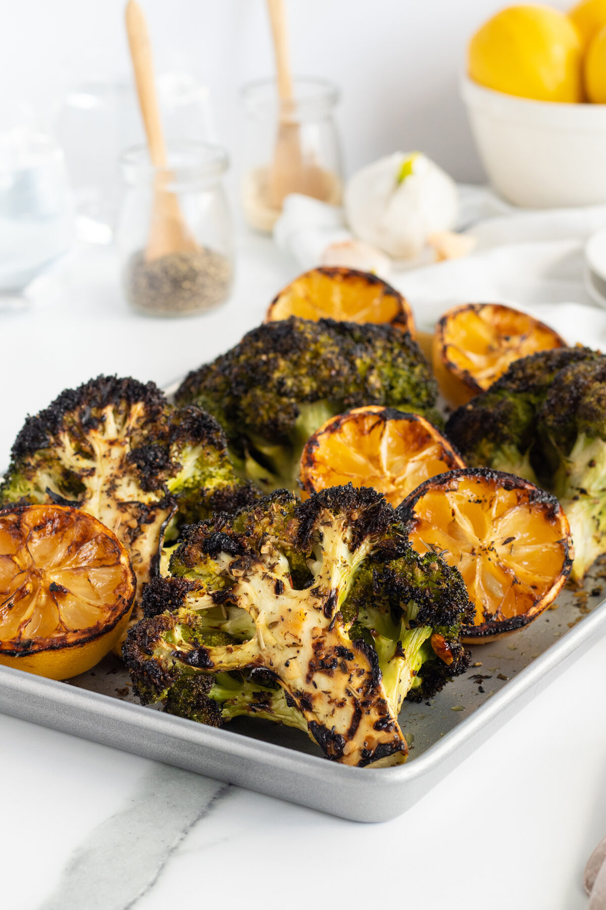 Roasted broccoli steaks and halved lemons with charred edges on a metal baking tray, set on a white kitchen surface with spices, garlic, and a bowl of lemons blurred in the background.