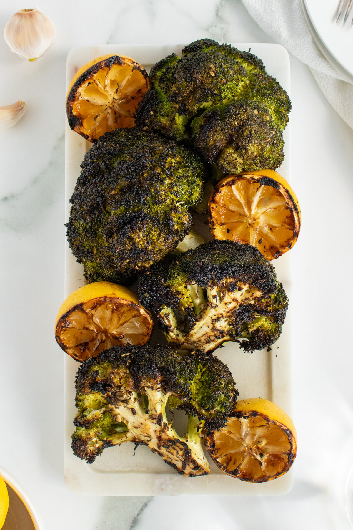 Charred roasted broccoli florets and halved grilled lemons arranged on a rectangular white serving platter, with a few garlic cloves nearby on a white marble surface.