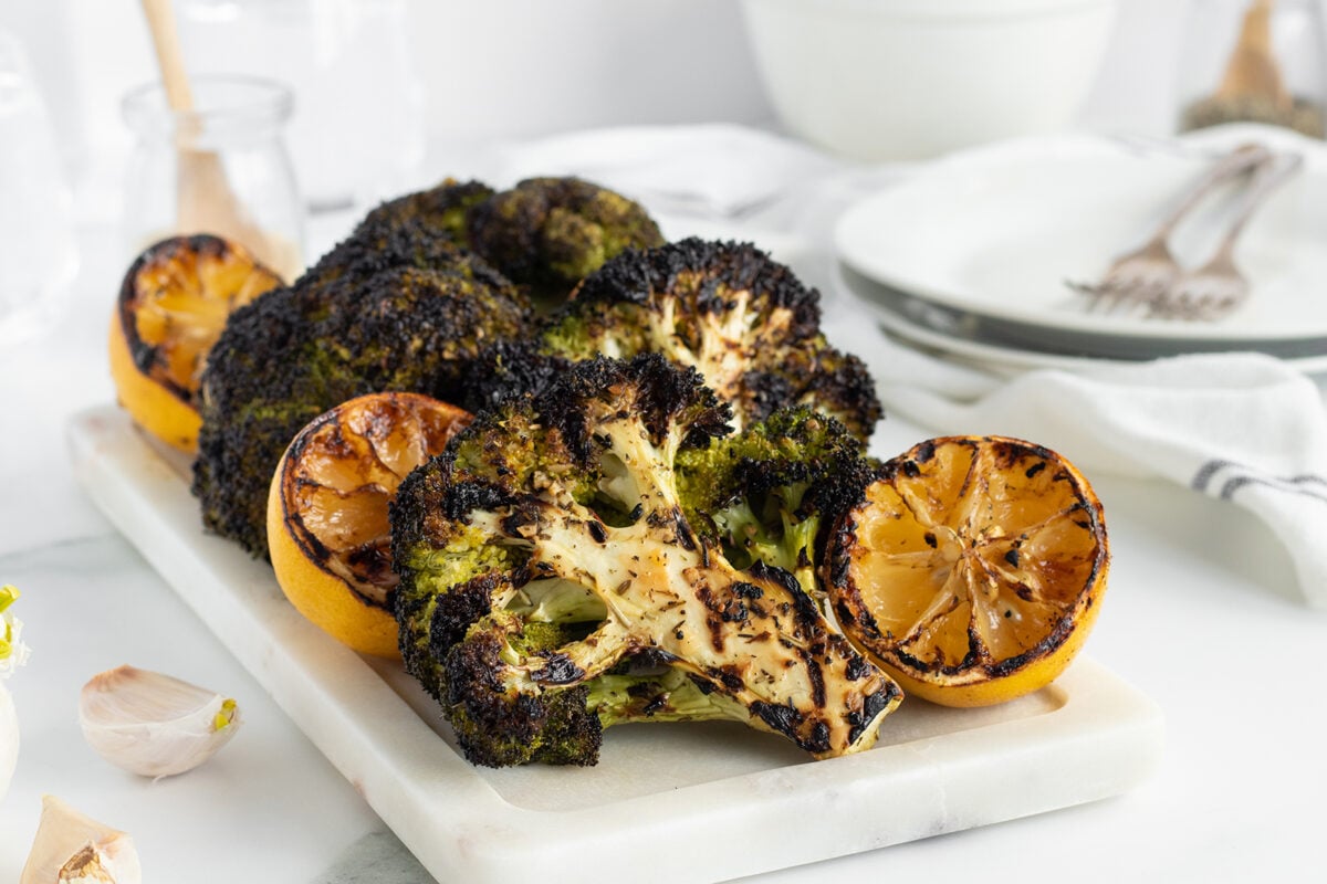 Charred broccoli florets and grilled lemon halves are arranged on a white marble serving board, set on a light table with plates, forks, garlic cloves, and a striped napkin in the background.