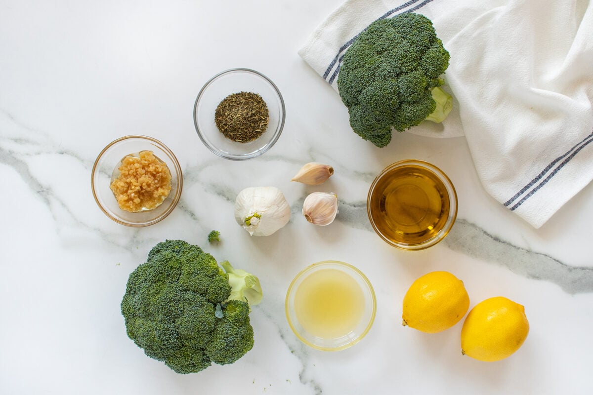 Broccoli heads, minced garlic, dried herbs, olive oil, garlic cloves, lemon juice, and whole lemons arranged on a white marble surface with a white towel.