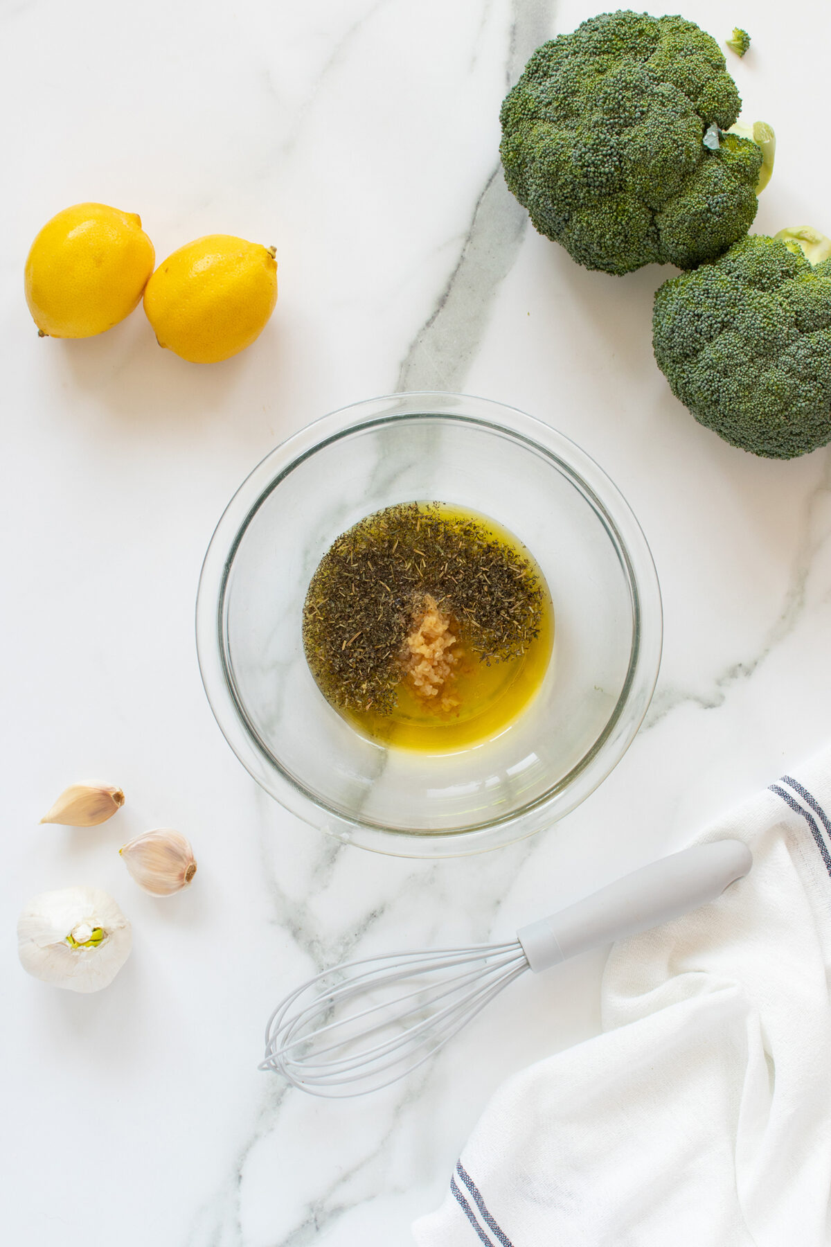 A glass bowl with olive oil, herbs, and minced garlic sits on a marble surface. Nearby are two lemons, two broccoli heads, garlic cloves, a whisk, and a white kitchen towel.