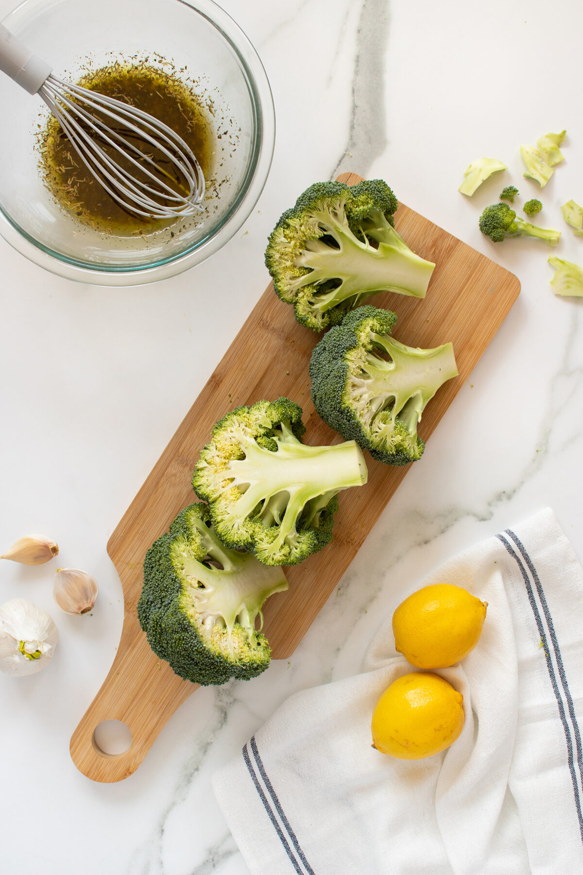 A cutting board with four broccoli halves, a whisk in a bowl of marinade, garlic cloves, lemon halves, and a white towel with blue stripes on a marble countertop.