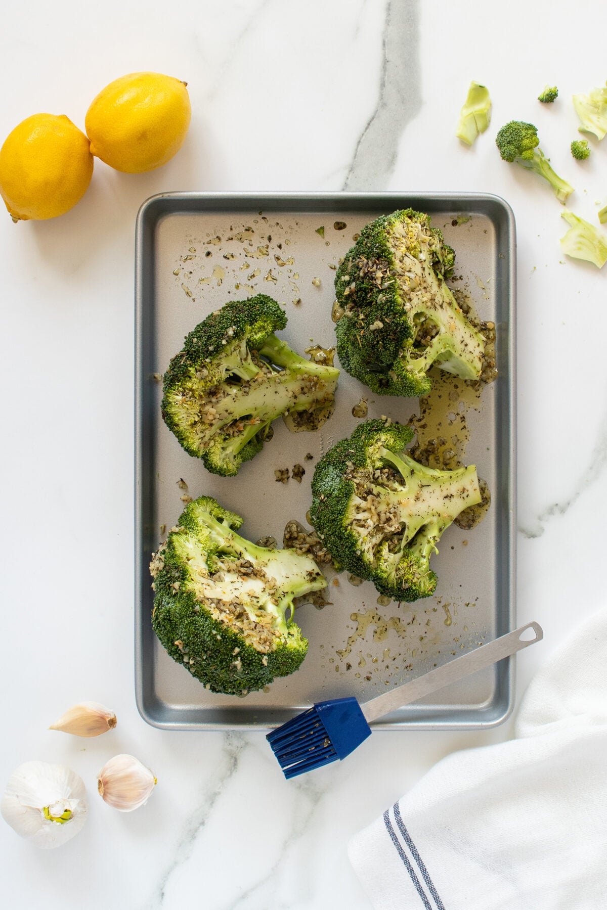 Four broccoli crowns on a baking sheet are seasoned with herbs. A blue brush rests on the tray. Nearby are two lemons, garlic cloves, and broccoli pieces on a white marble countertop with a white and blue towel.