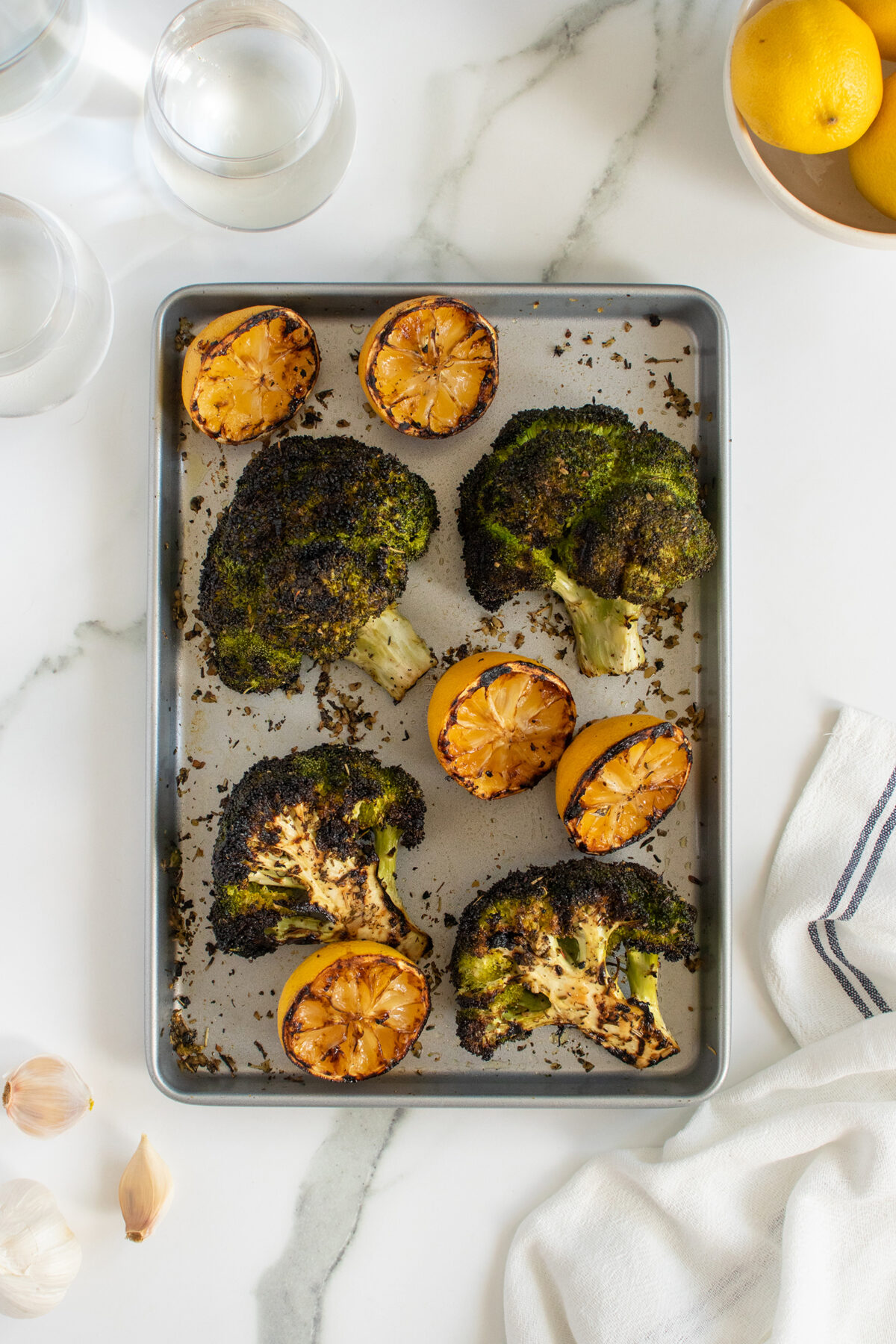 A baking sheet with roasted broccoli florets and halved lemons, set on a white marble surface with two glasses of water, whole lemons, garlic cloves, and a striped napkin nearby.