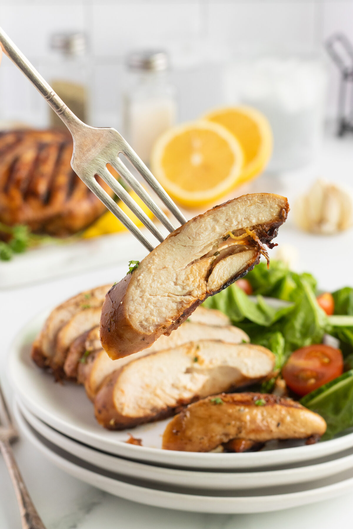 A fork holds a juicy slice of grilled chicken breast above a plate with more chicken slices, fresh spinach, and cherry tomatoes. Sliced oranges and seasonings are visible in the background.