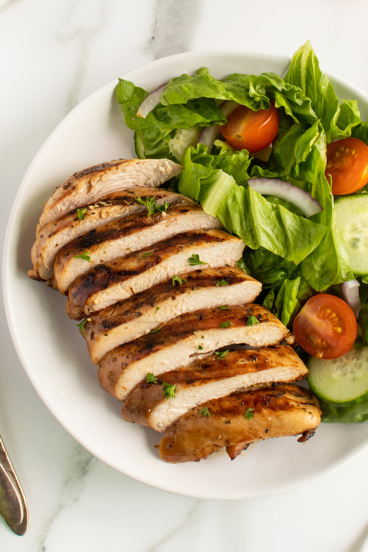 A white plate with sliced grilled chicken breast, garnished with herbs, served alongside a fresh salad of leafy greens, cherry tomatoes, cucumber slices, and red onion.