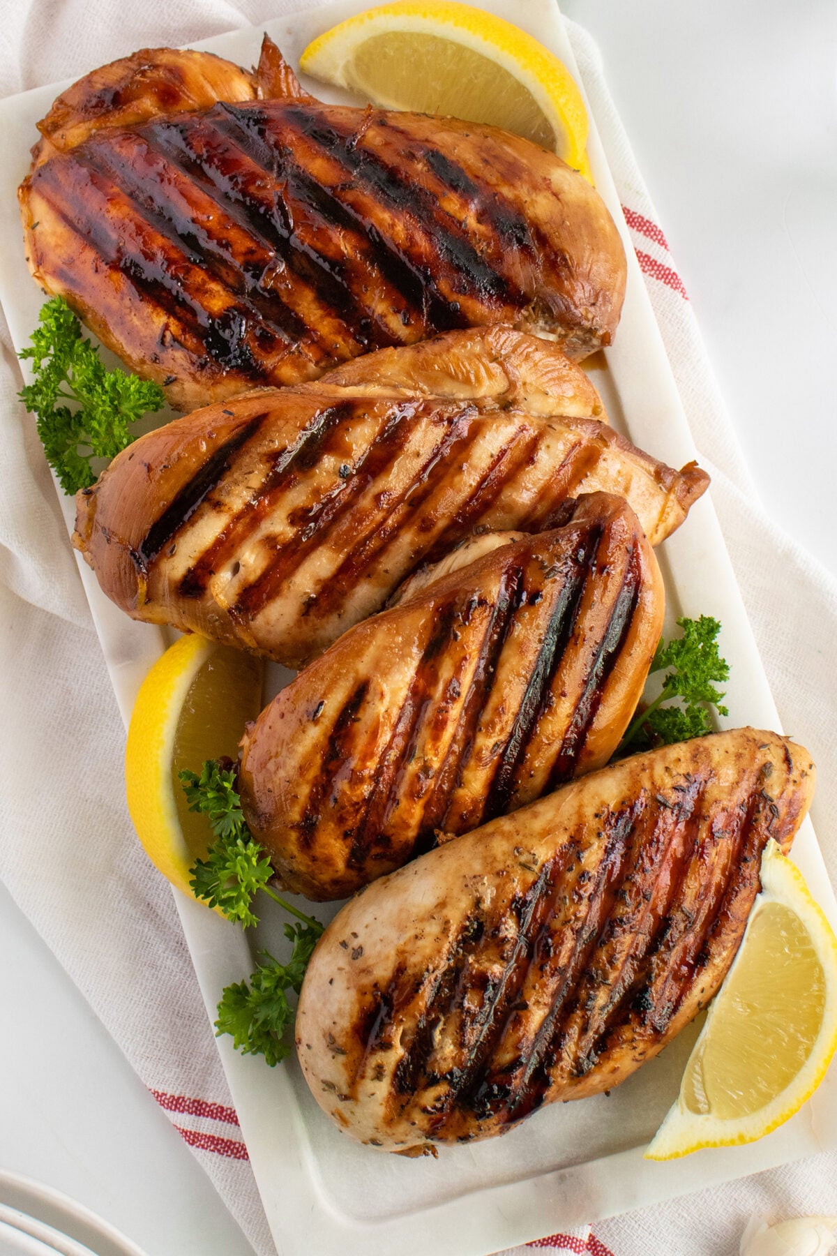Four grilled chicken breasts with visible grill marks are arranged on a white rectangular plate, garnished with parsley and lemon wedges, atop a red-striped white cloth.