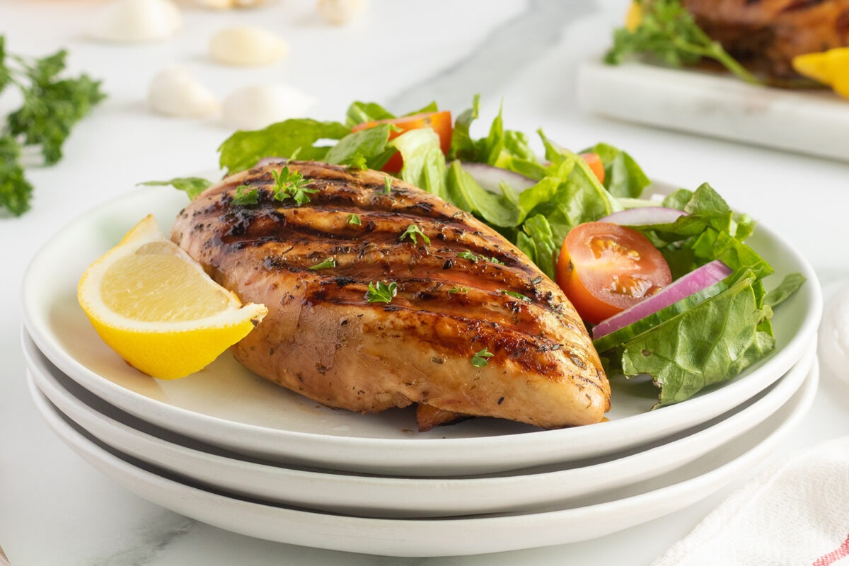 Grilled chicken breast garnished with herbs, served with a fresh salad of lettuce, tomatoes, and red onions, and a lemon wedge on the side, on a white plate.