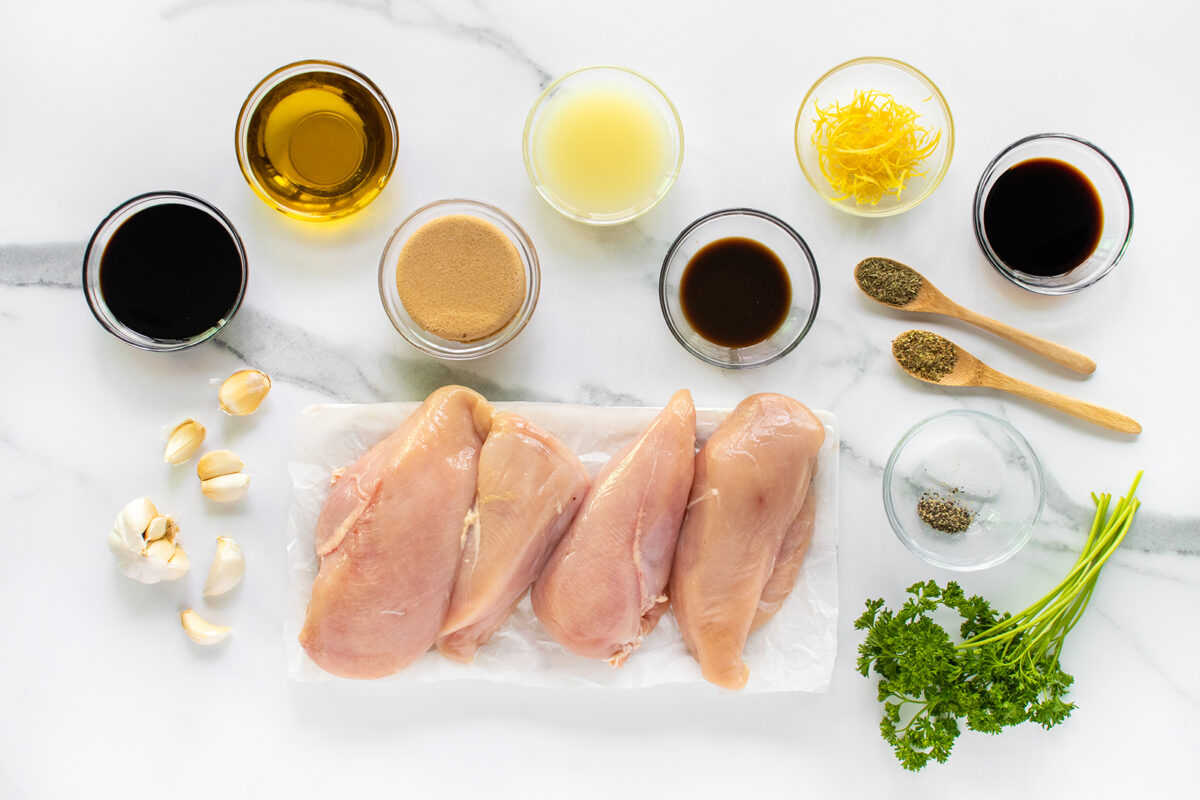 Raw chicken breasts on parchment with parsley, black pepper, garlic cloves, dried herbs, brown sugar, soy sauce, olive oil, lemon juice, lemon zest, and balsamic vinegar in small bowls on a white marble surface.