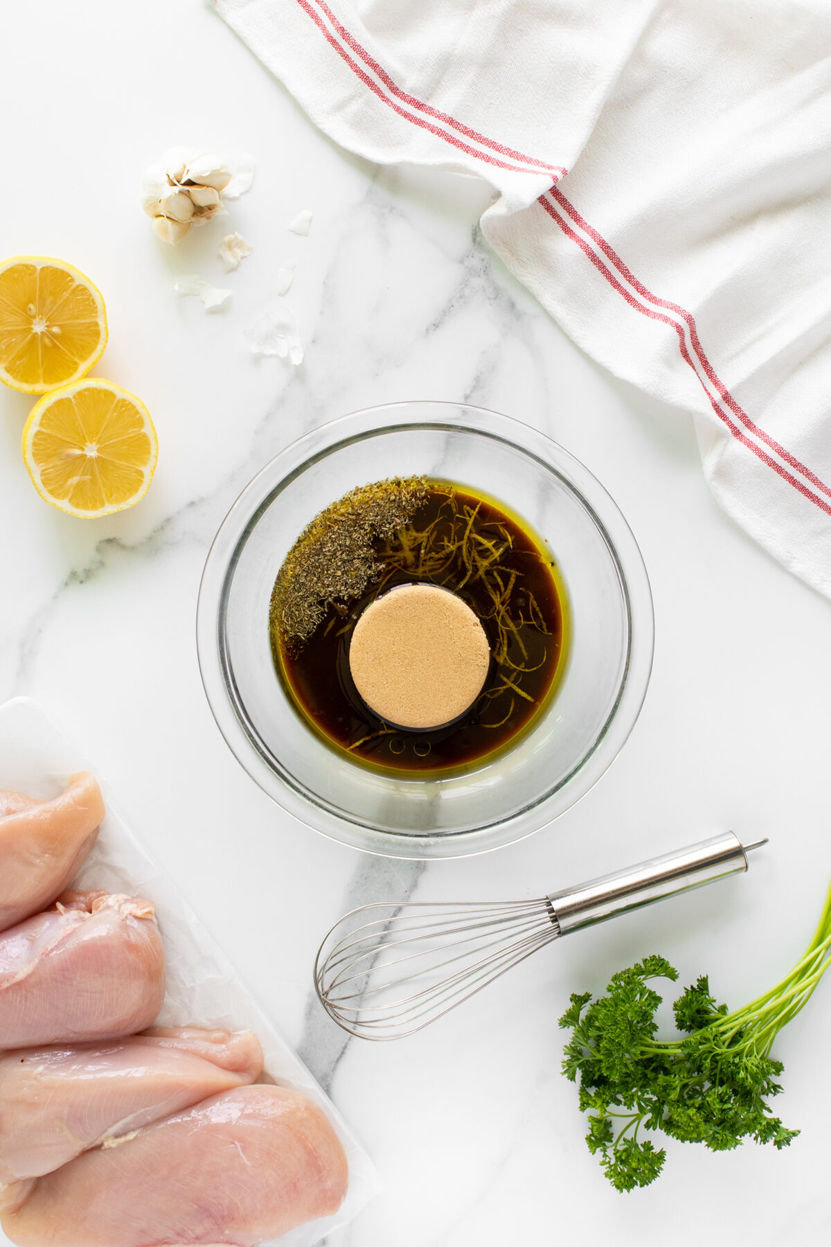 A glass bowl with marinade ingredients, a whisk, raw chicken breasts, lemon halves, garlic, parsley, and a white kitchen towel on a marble surface.