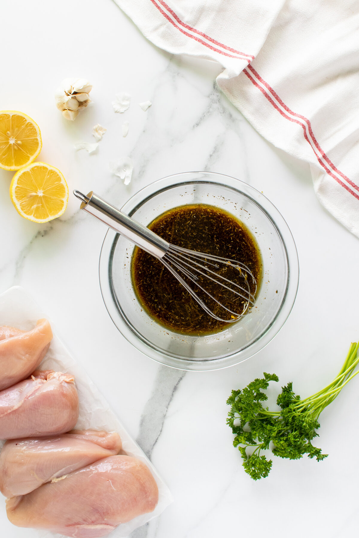 A glass bowl of marinade with a whisk, raw chicken breasts, halved lemons, parsley, garlic cloves, and a white towel with a red stripe on a white marble surface.