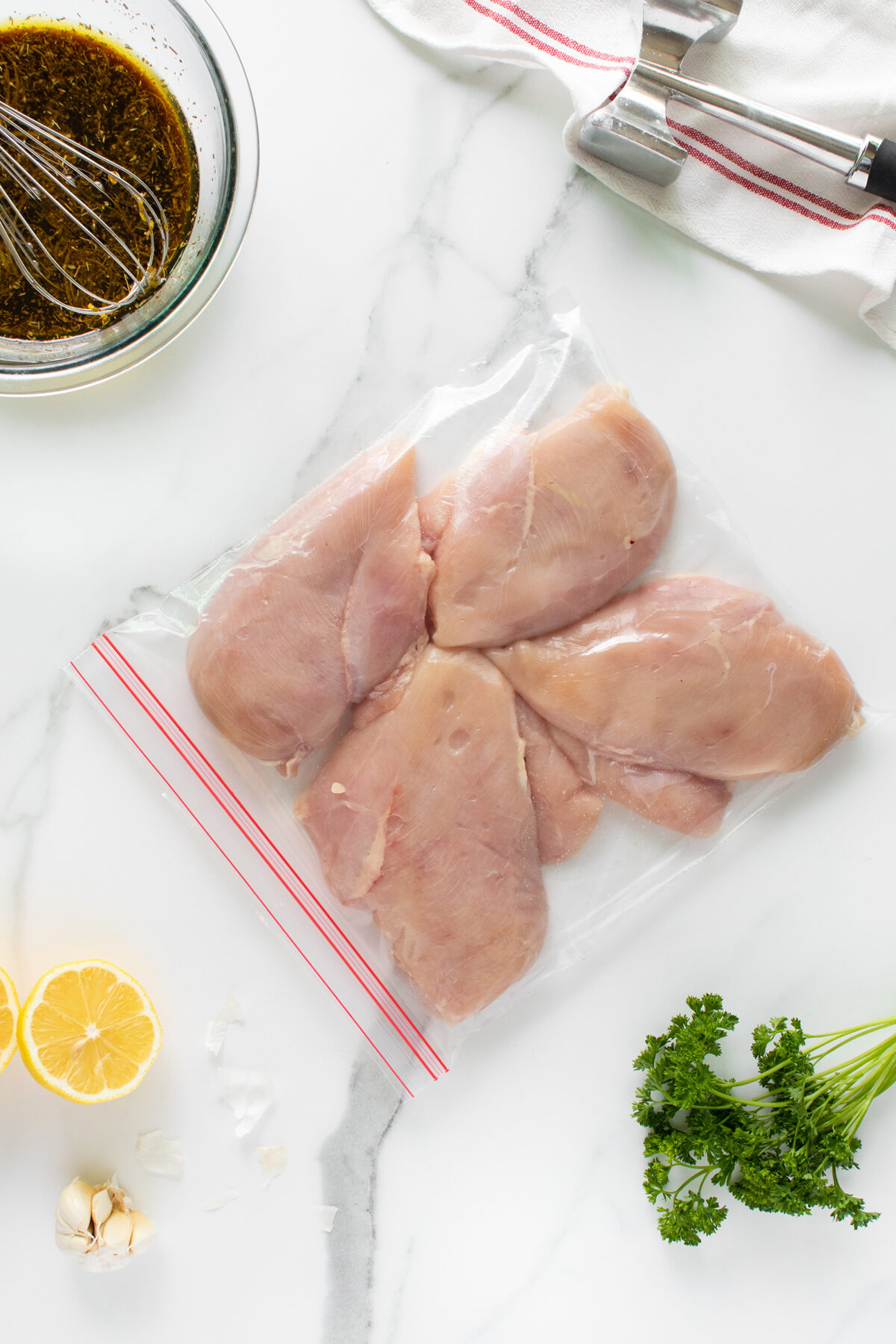 Raw chicken breasts in a resealable plastic bag on a marble surface, with lemon halves, fresh parsley, garlic cloves, a meat tenderizer, and a bowl of marinade visible nearby.