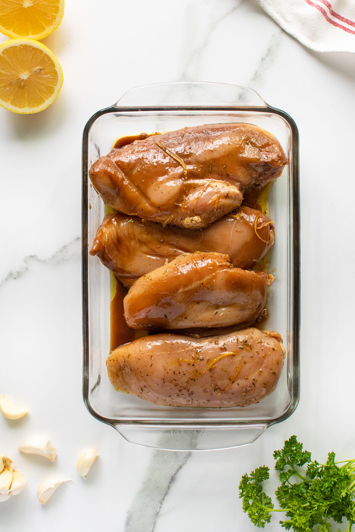 Four raw chicken breasts marinating in sauce in a glass baking dish on a white marble surface. Fresh garlic, lemon halves, parsley, and a striped towel are nearby.