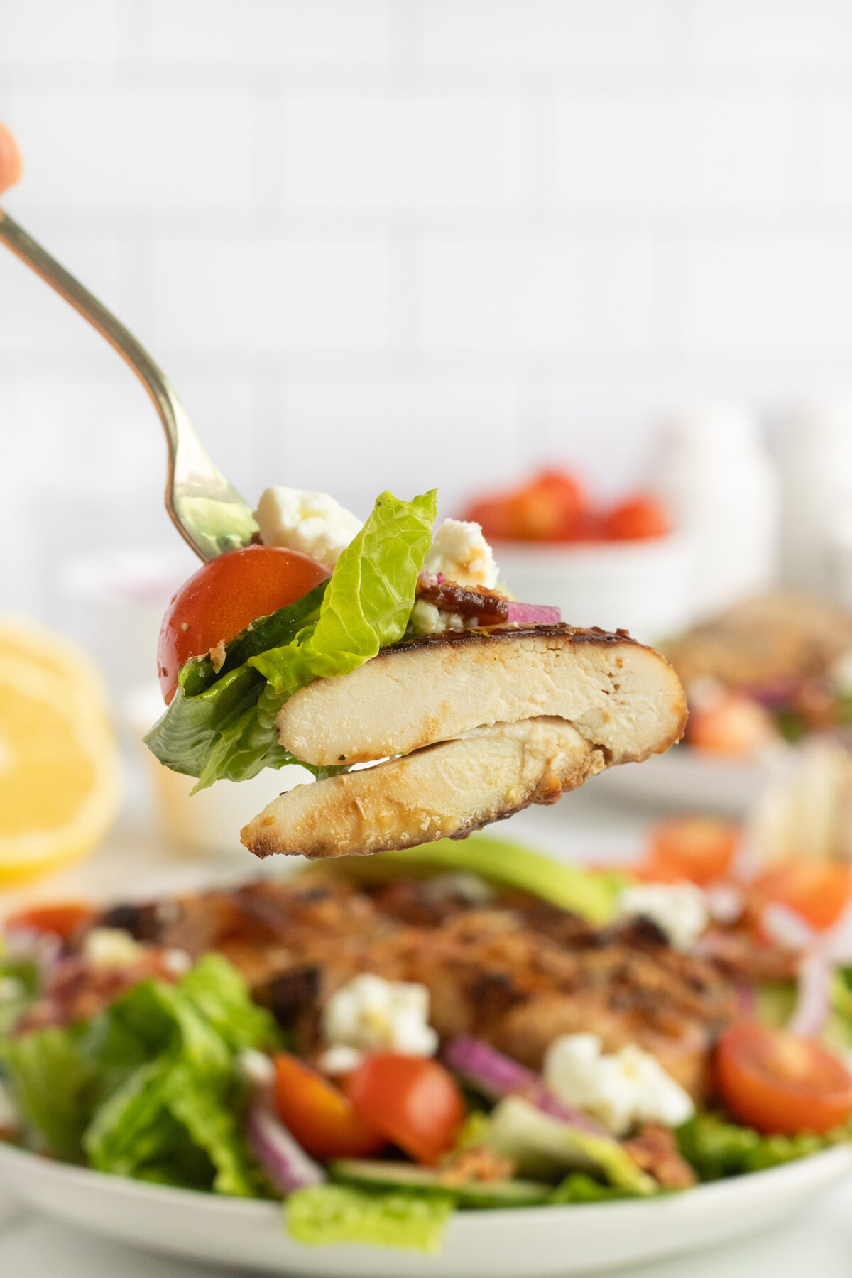 A fork holds a bite of salad with grilled chicken, lettuce, cherry tomato, red onion, and feta cheese above a plate of similar salad in the background.