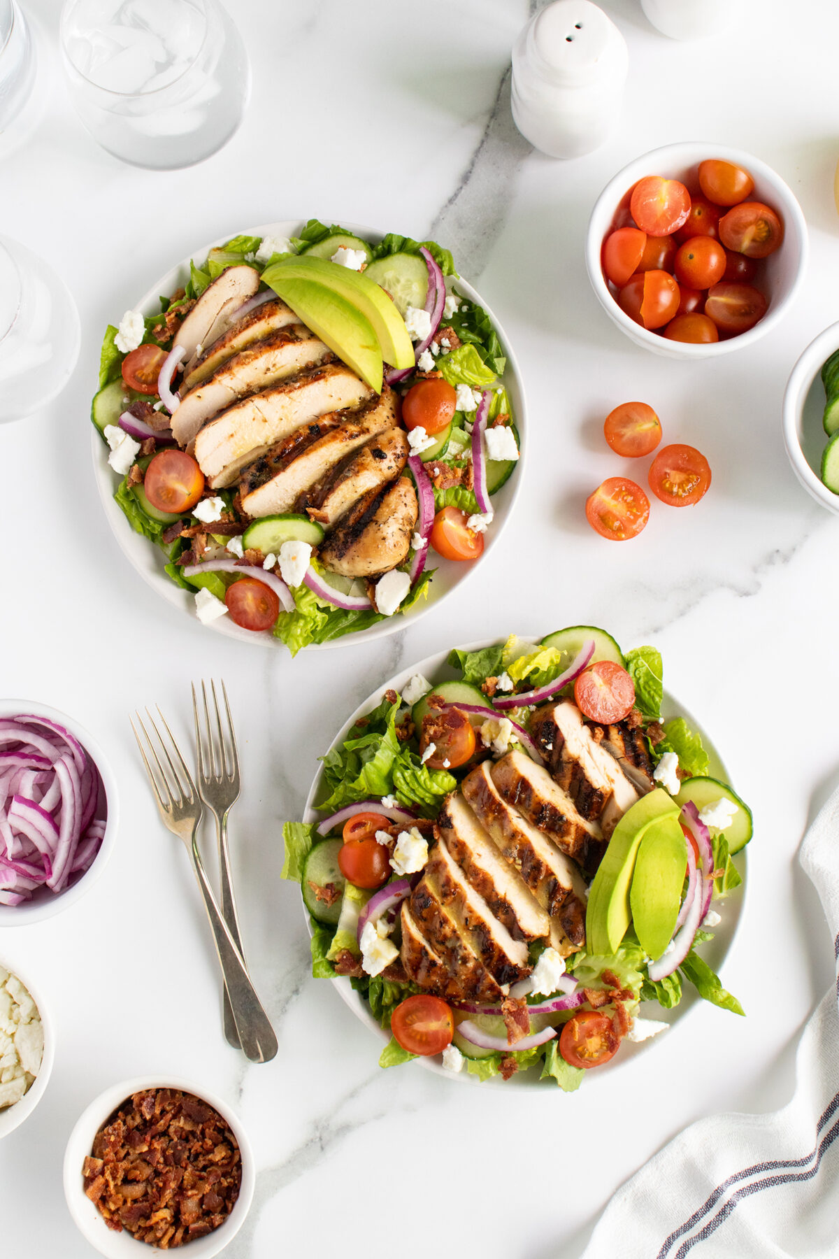 Two plates of grilled chicken salad with avocado, cherry tomatoes, red onion, and feta cheese on a white surface. Small bowls of cherry tomatoes, cucumber, red onion, bacon bits, and feta are nearby. Two forks rest beside the plates.
